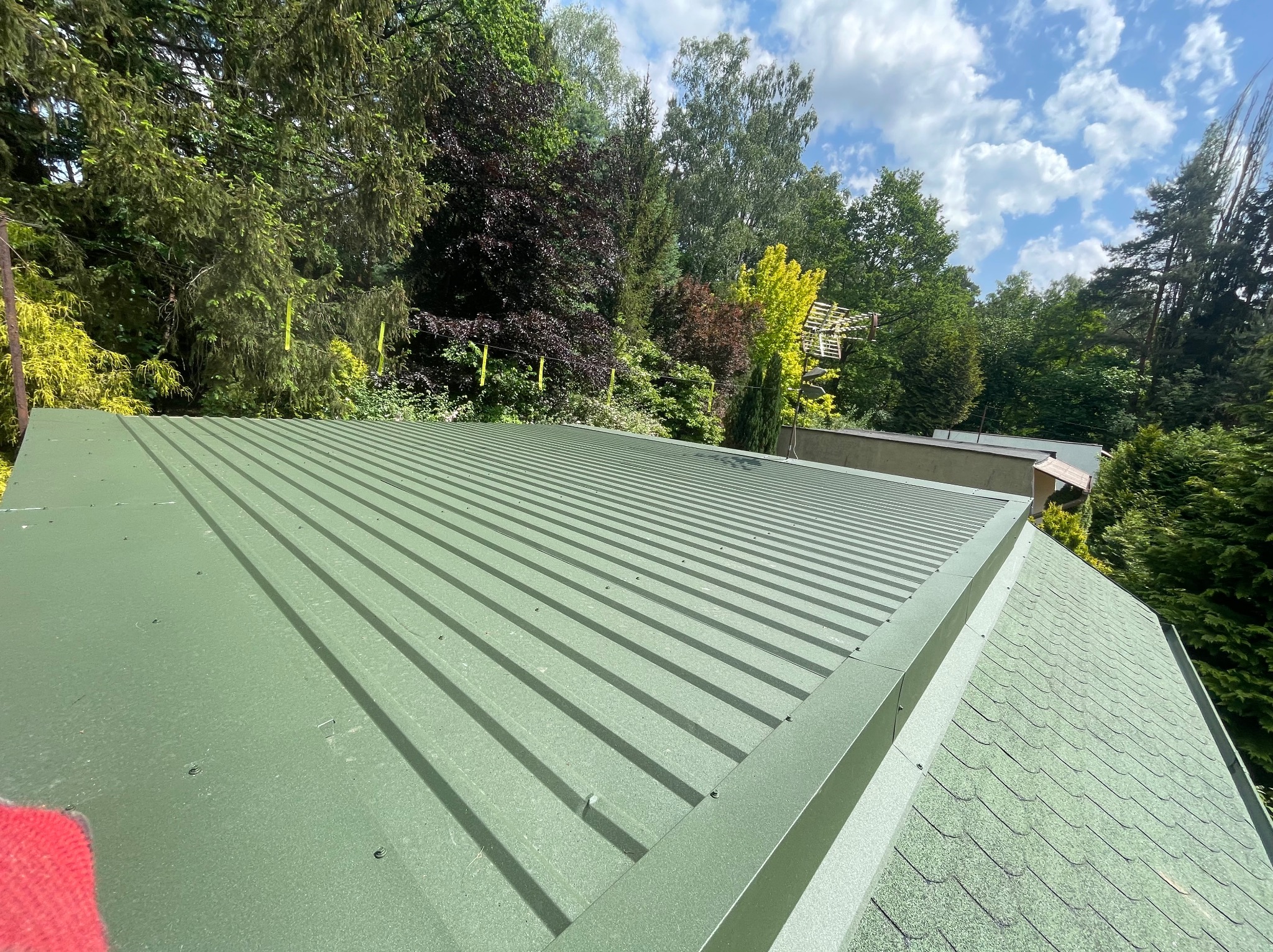 Zielony dach z blachy trapezowej na tle drzew i błękitnego nieba z chmurami, widok z góry.