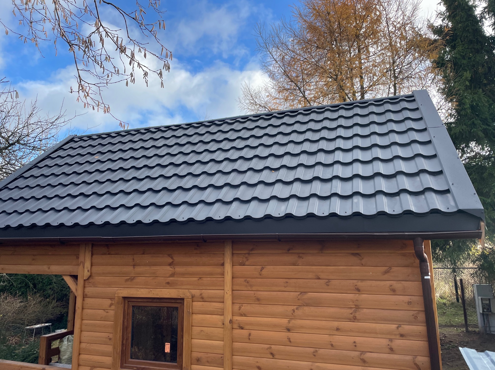 Nowy, grafitowy dach z blachy trapezowej na drewnianym domku letniskowym, widok z dołu na tle błękitnego nieba z chmurami i jesiennych drzew.
