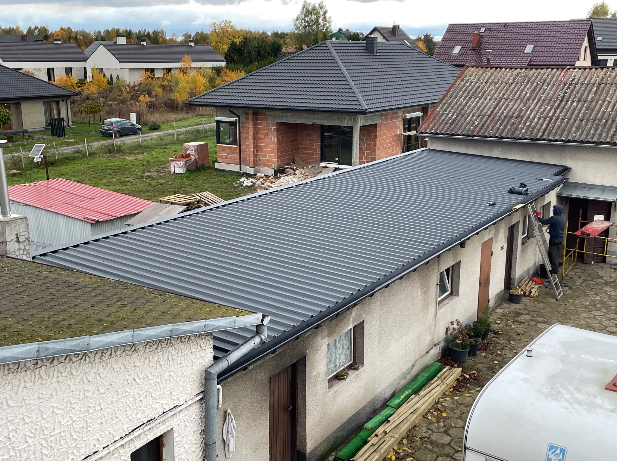Szary dach z blachy trapezowej na budynku gospodarczym podczas montażu, widoczna drabina i pracownik w ciemnej odzieży, w tle domy jednorodzinne i jesienne drzewa.