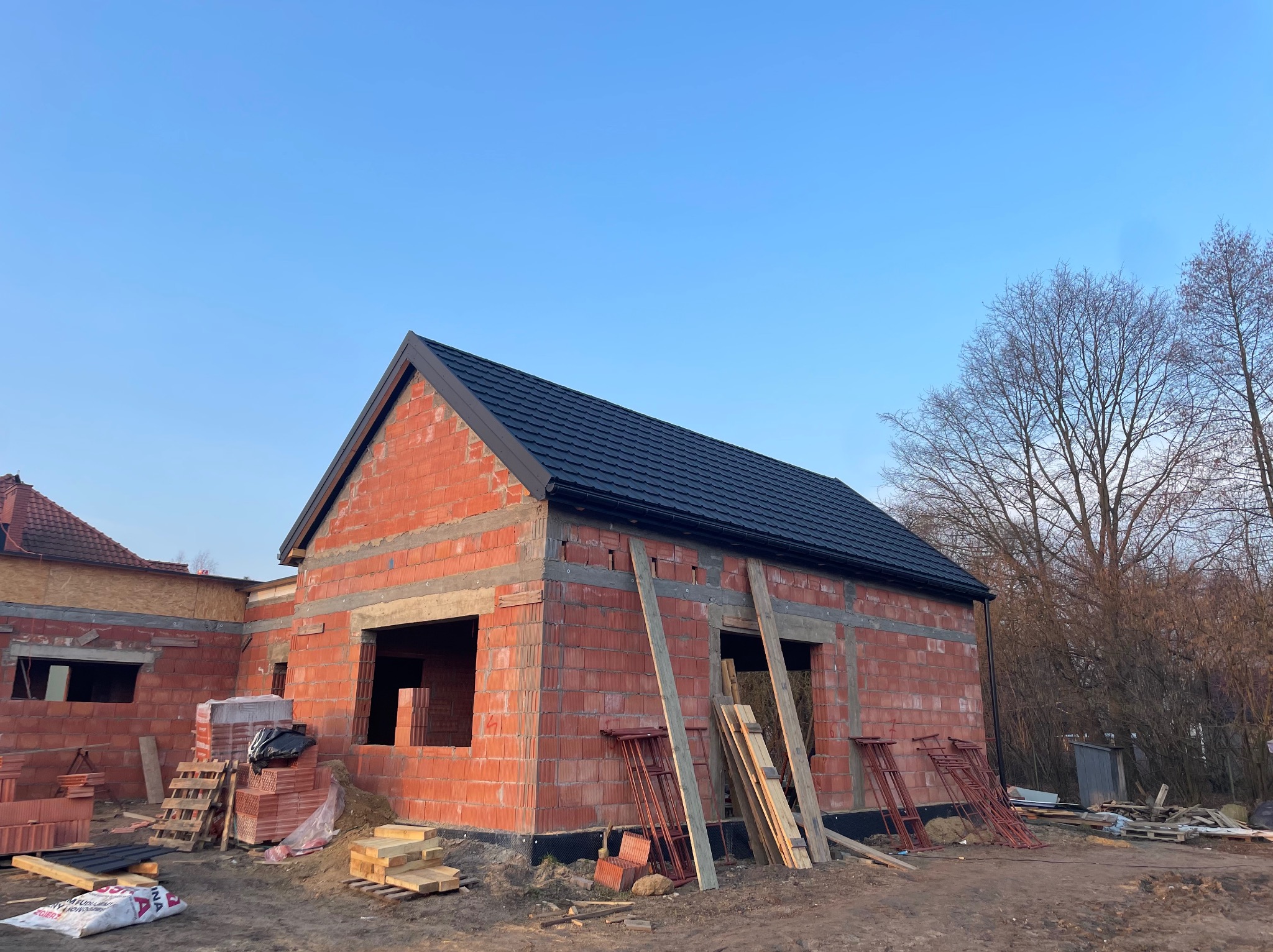Surowy budynek z czerwonej cegły w trakcie budowy, z zamontowaną czarną rynną i fragmentem ciemnego pokrycia dachowego, na tle błękitnego nieba i drzew bez liści.