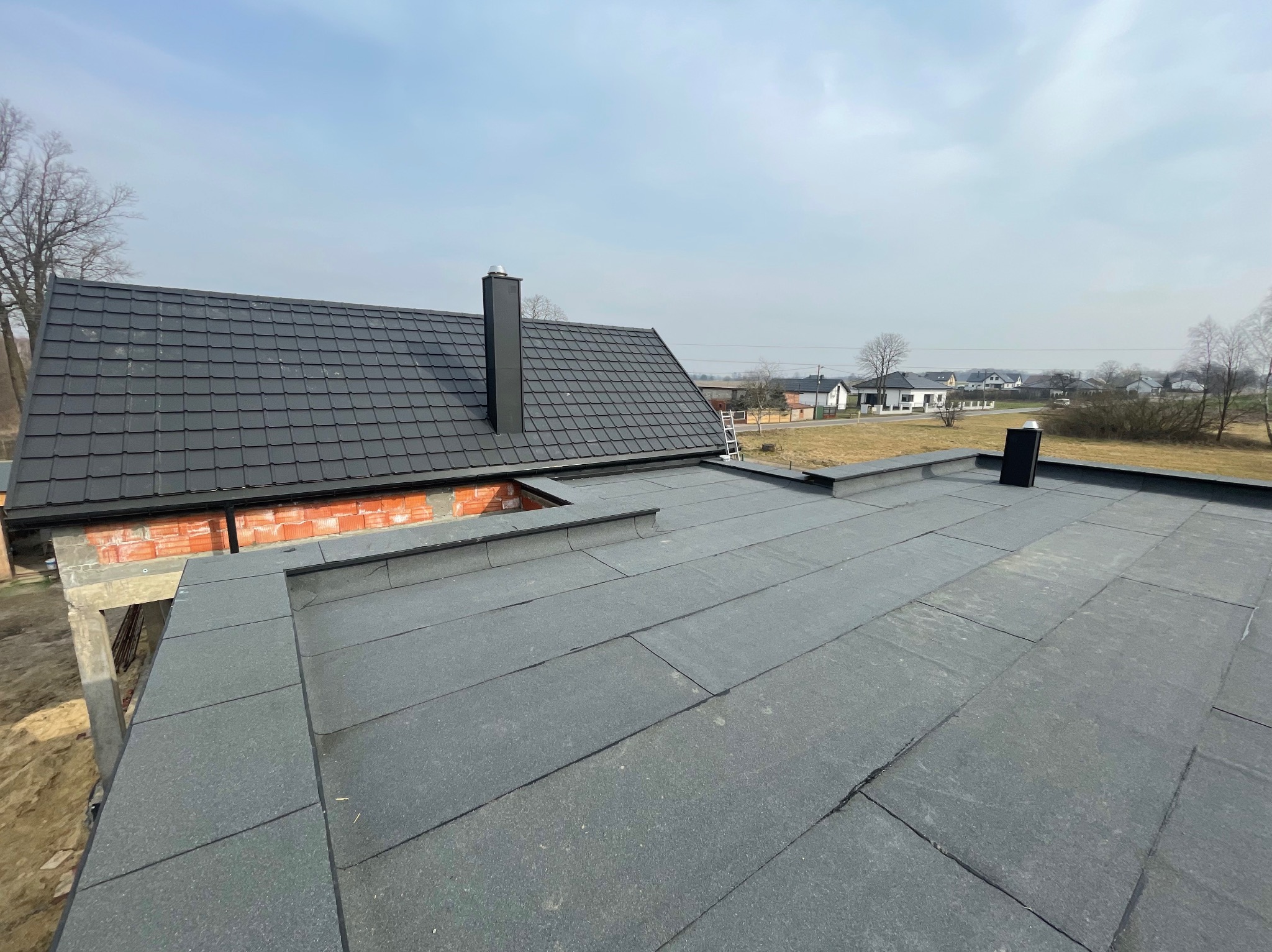 Widok z góry na fragment dachu pokrytego czarną papą bitumiczną na płasko, z kominem i fragmentem dachu skośnego z czarnej dachówki modułowej, w tle budynki mieszkalne.