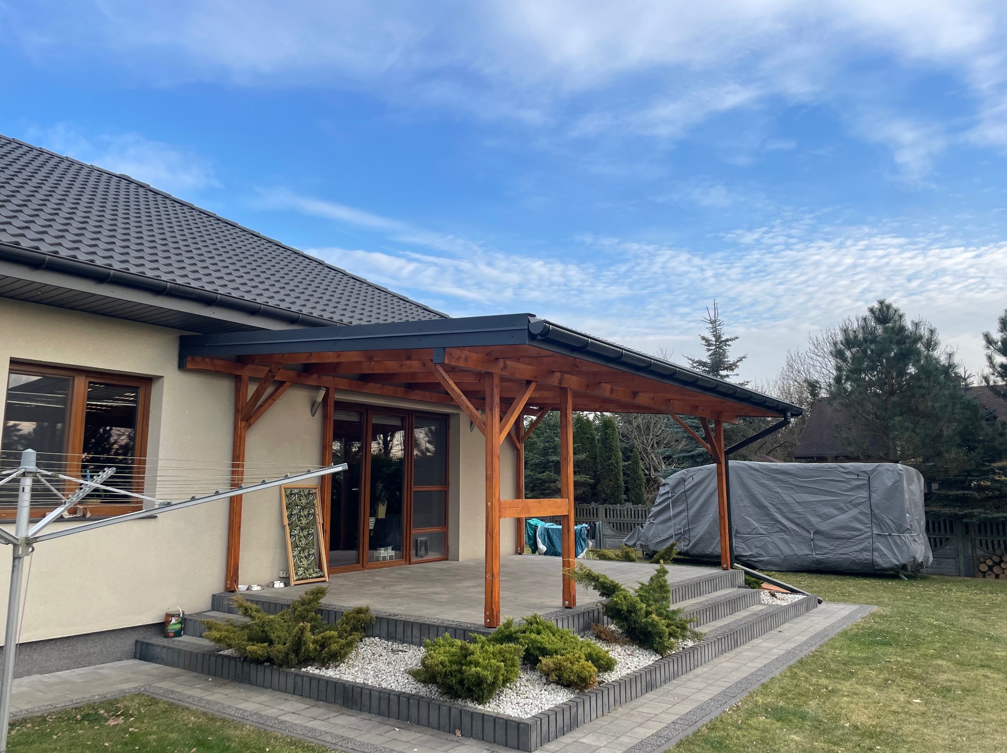 Zadaszony taras z drewnianą konstrukcją przylegający do domu z szarym dachem i rynną, widoczne schody z kostki i rośliny ozdobne.