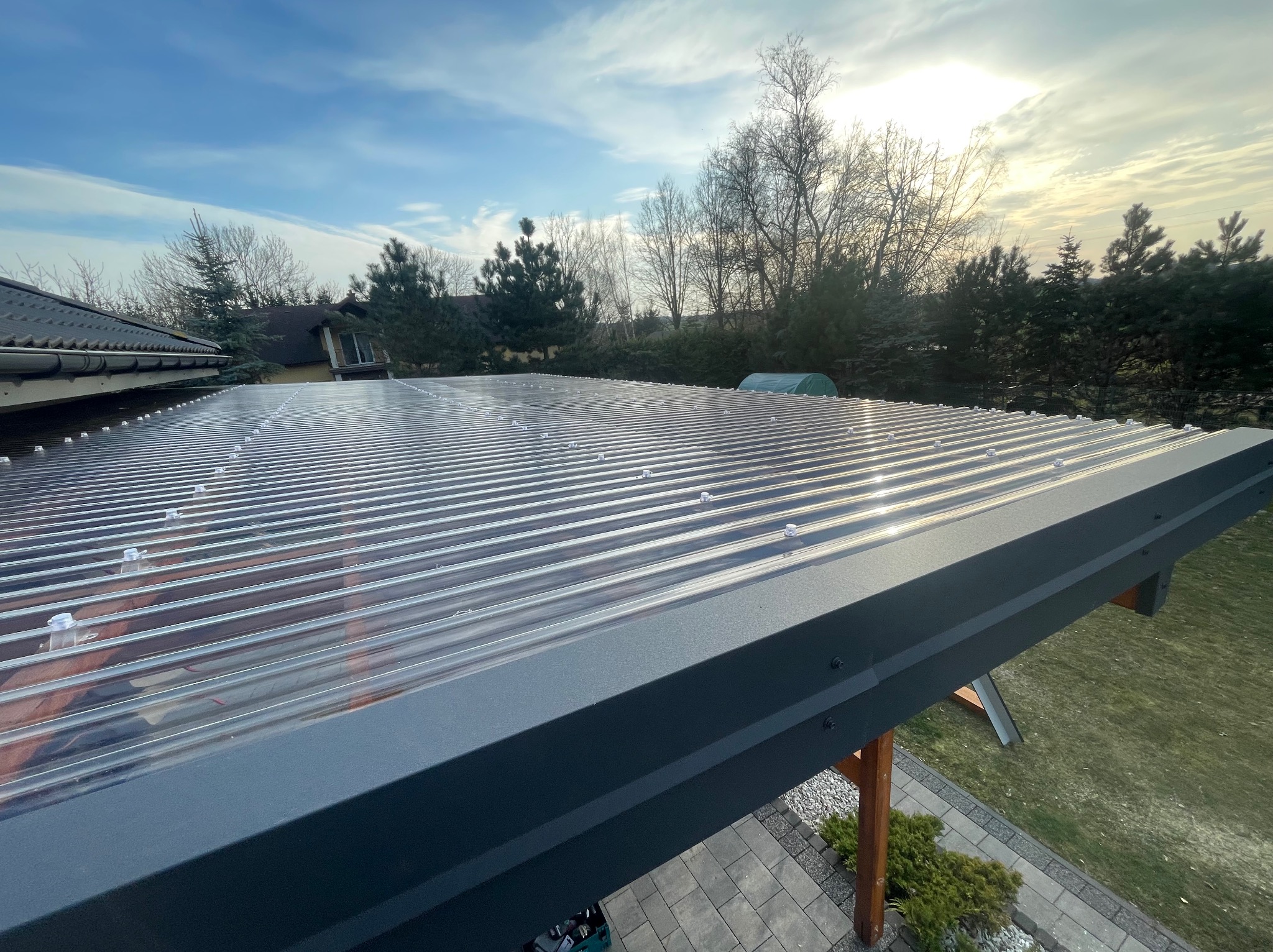 Przezroczysty dach z blachy trapezowej, mocowany śrubami, z widoczną konstrukcją wspornikową na tle ogrodu i błękitnego nieba.