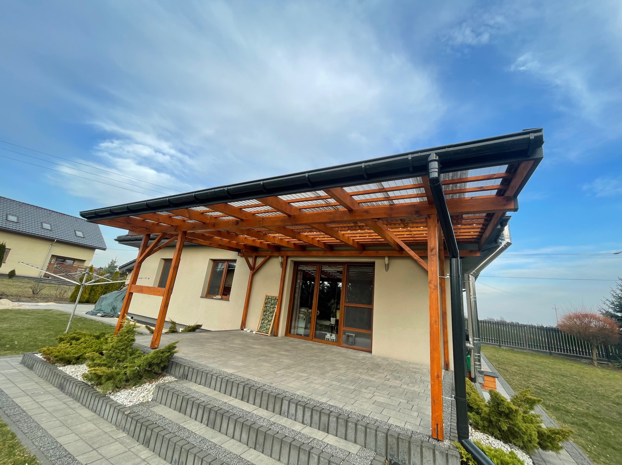 Drewniana pergola z przezroczystym zadaszeniem przylegająca do domu, widoczne elementy konstrukcyjne, rynna i schody z kostki brukowej.