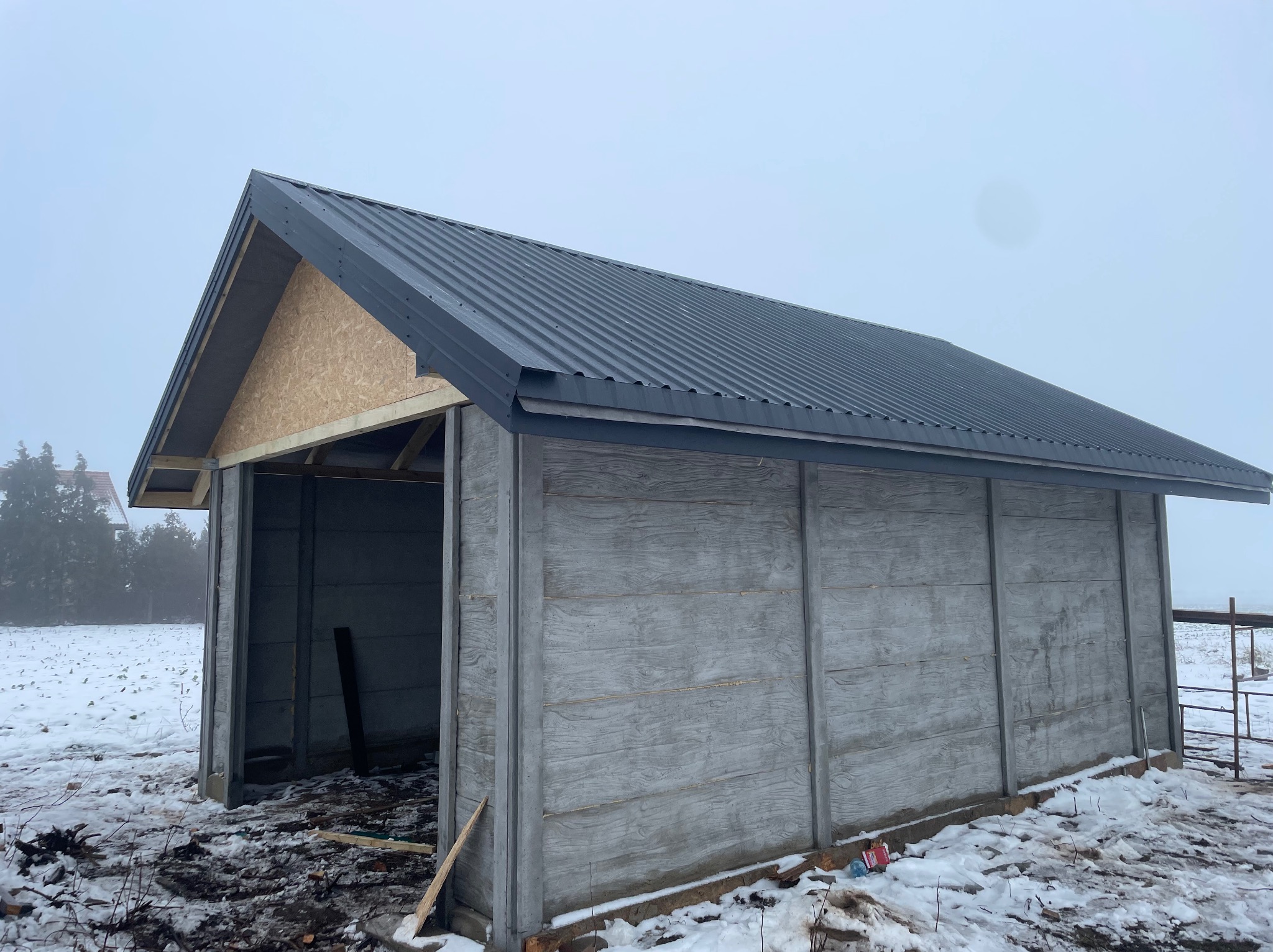Konstrukcja wiaty z prefabrykatów betonowych i blachy trapezowej na dachu, w zimowej scenerii, z widoczną niezabudowaną ścianą szczytową z płyty OSB.