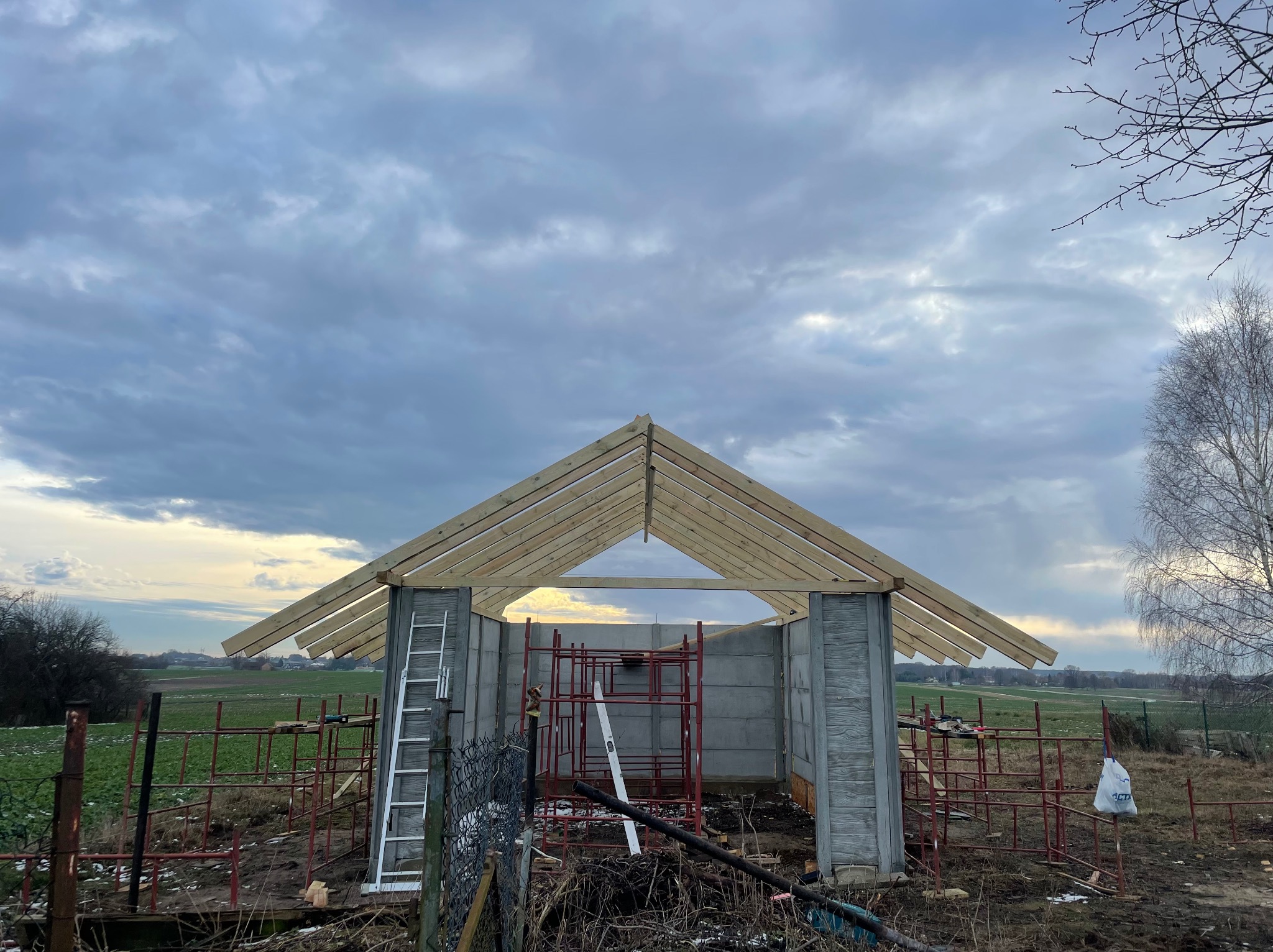 Konstrukcja małego budynku z betonu komórkowego w trakcie budowy, widoczna więźba dachowa z surowego drewna, rusztowania i drabina, krajobraz polny w tle pod pochmurnym niebem.