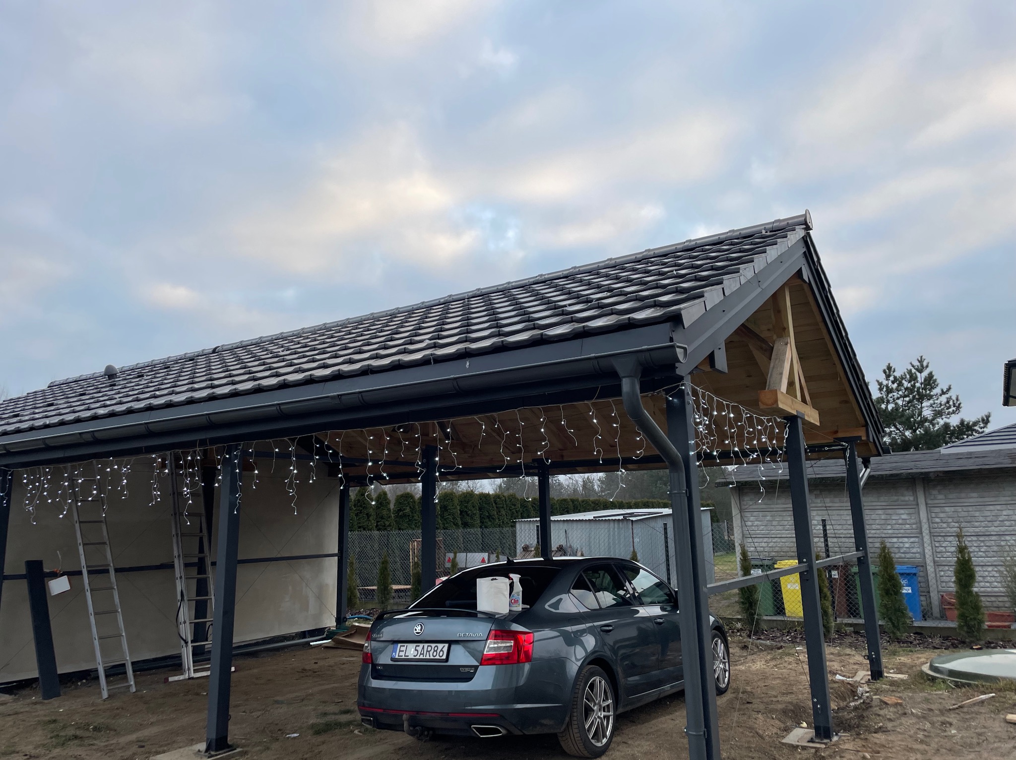 Szara Skoda Octavia pod zadaszeniem z ciemną dachówką, ozdobionym lampkami, na tle pochmurnego nieba i ogrodzenia.