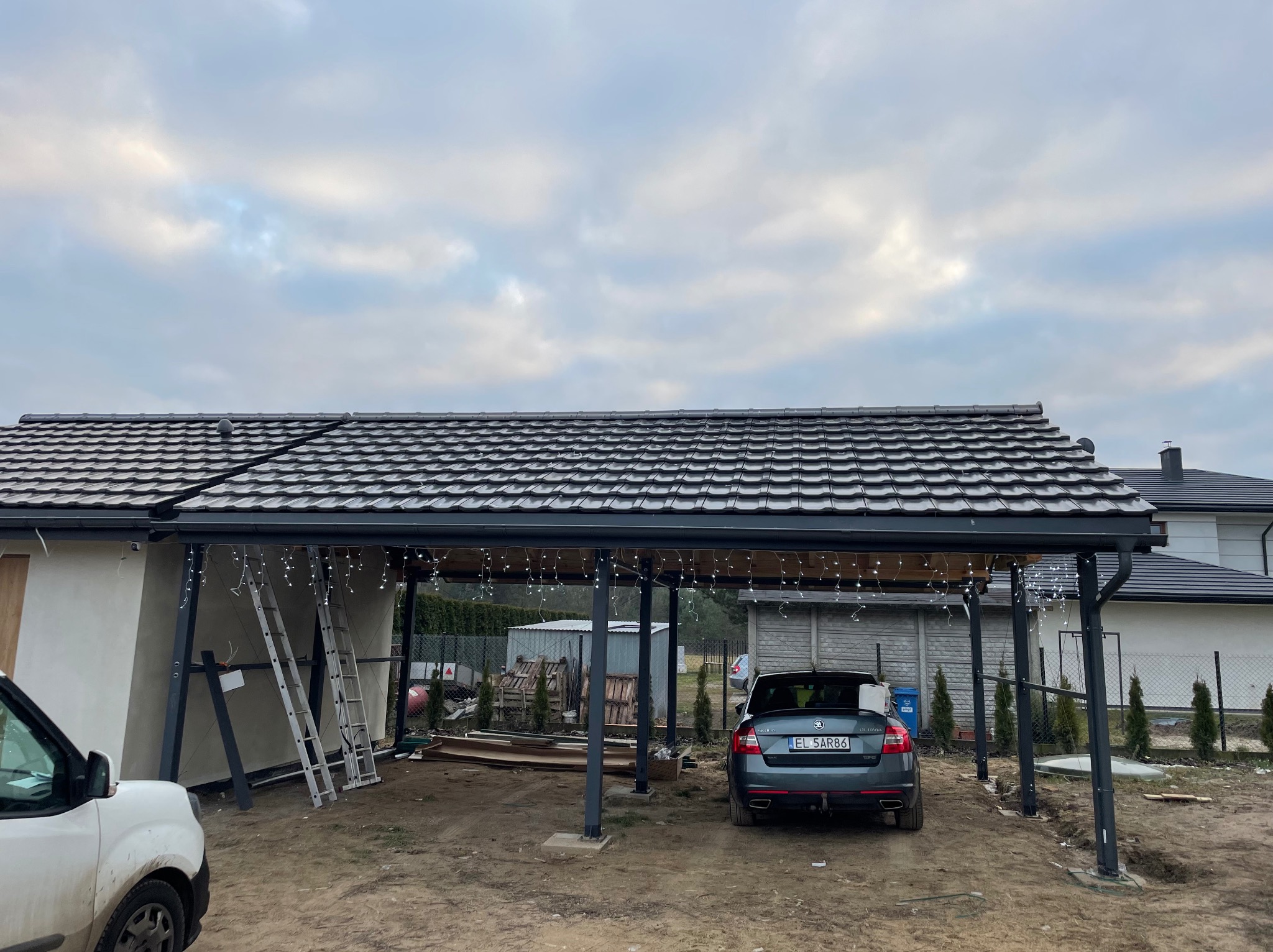 Zadaszenie posesji z ciemną dachówką karpiówką, oświetlone girlandami, z samochodem na podjeździe i drabiny opartymi o ścianę budynku w tle pochmurne niebo.