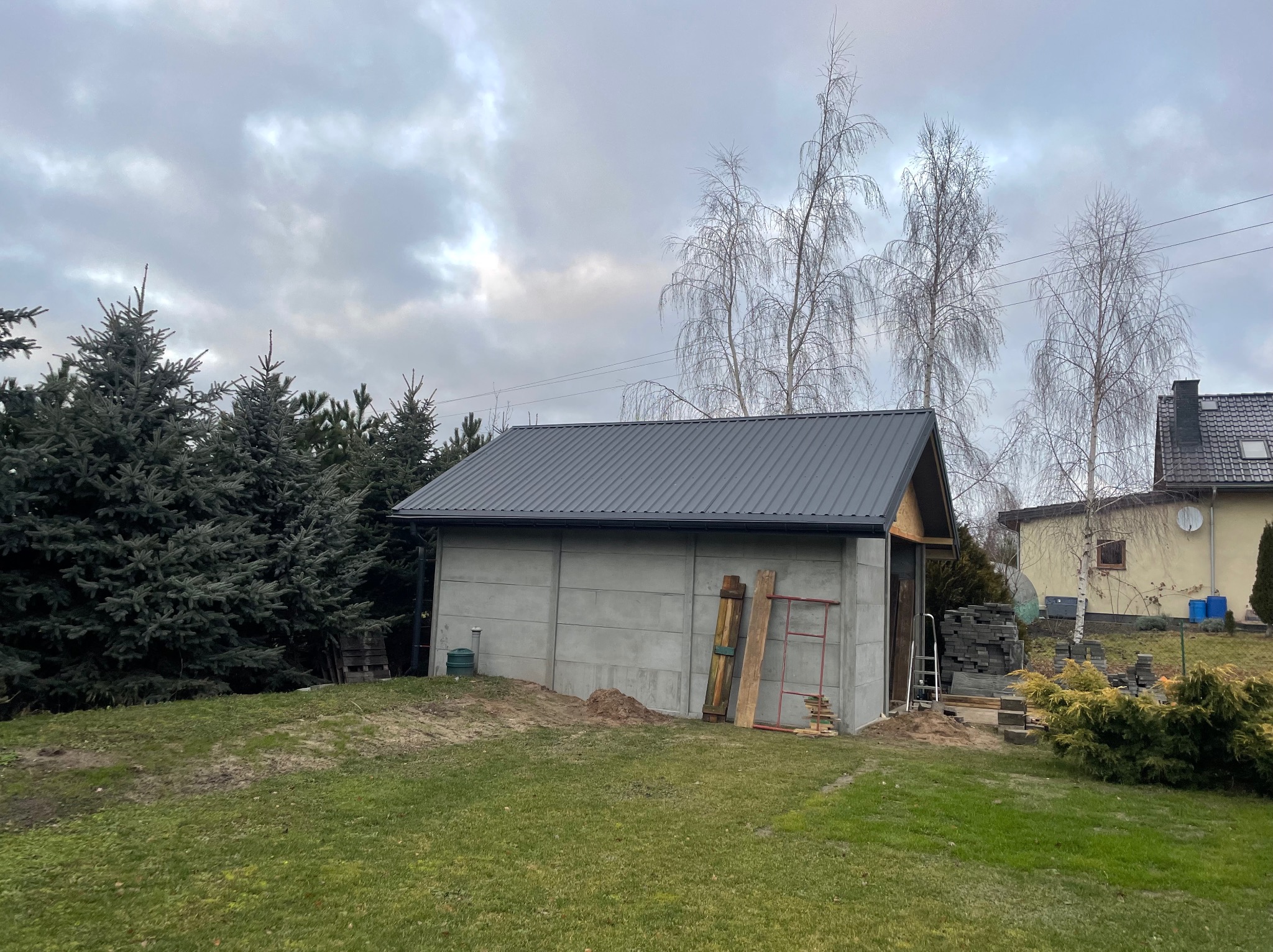 Nowoczesny, szary budynek gospodarczy z blachy trapezowej na dachu, widoczny w ogrodzie z drzewami iglastymi i brzozami na tle pochmurnego nieba.