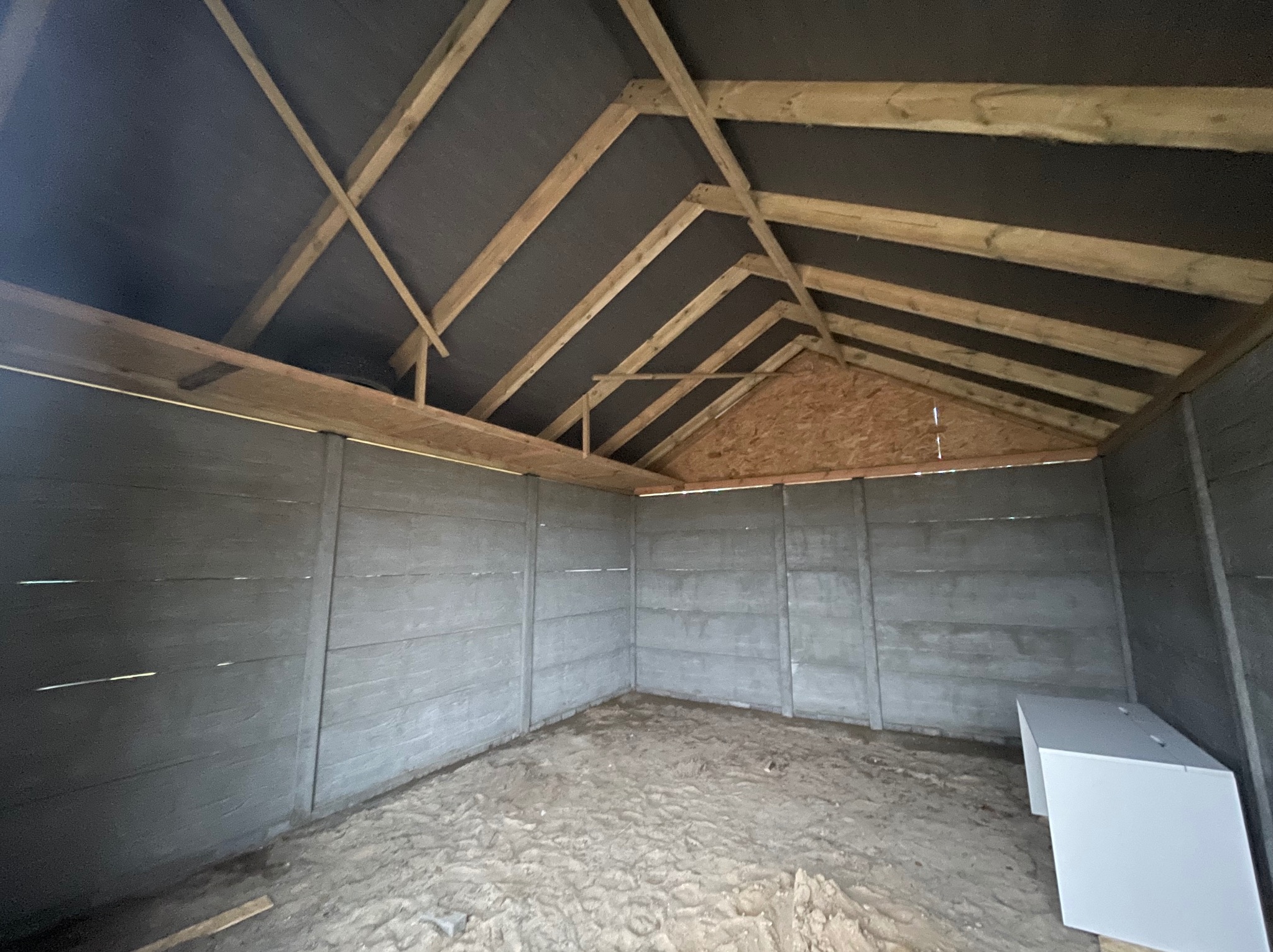 Wnętrze nowo wzniesionej konstrukcji z drewnianą więźbą dachową pokrytą czarną membraną, szare betonowe ściany z widocznymi łączeniami, piaszczysta podłoga i biała szafka w rogu.