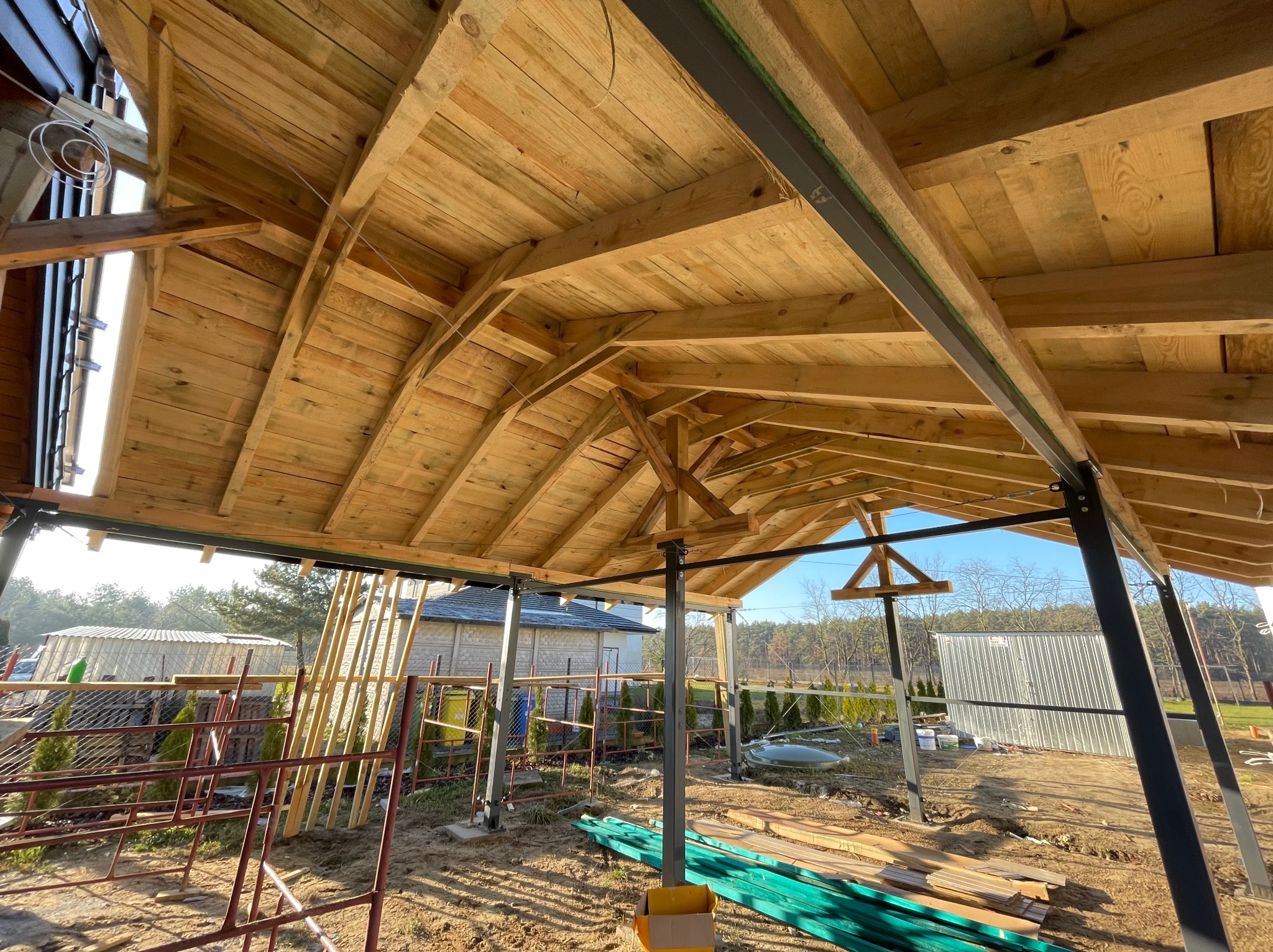 Wnętrze nowo budowanego drewnianego zadaszenia z widoczną konstrukcją więźby dachowej i metalowymi słupami nośnymi, w tle zabudowa mieszkalna i drzewa.