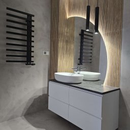 Artis Kuchnie na wymiar - Nowoczesna łazienka z owalną umywalką nablatową, szafką z szufladami, grzejnikiem drabinkowym w kolorze czarnym, podświetlanym lustrem w kształcie łuku i drewnianą ścianą z lameli.