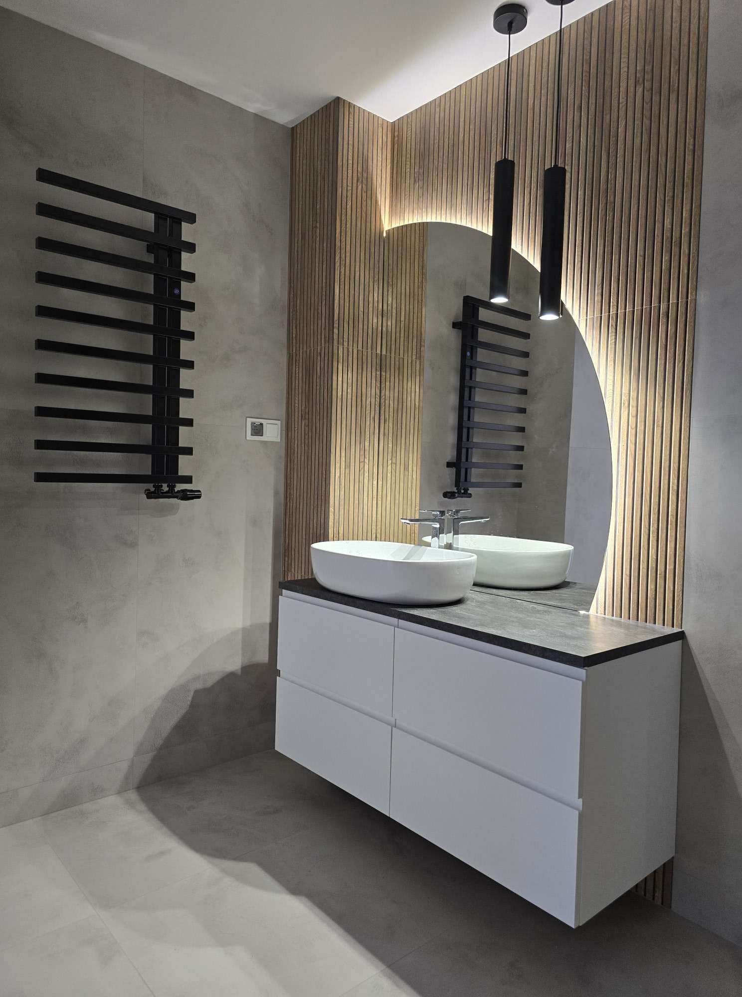 Nowoczesna łazienka z owalną umywalką nablatową, szafką z szufladami, grzejnikiem drabinkowym w kolorze czarnym, podświetlanym lustrem w kształcie łuku i drewnianą ścianą z lameli.