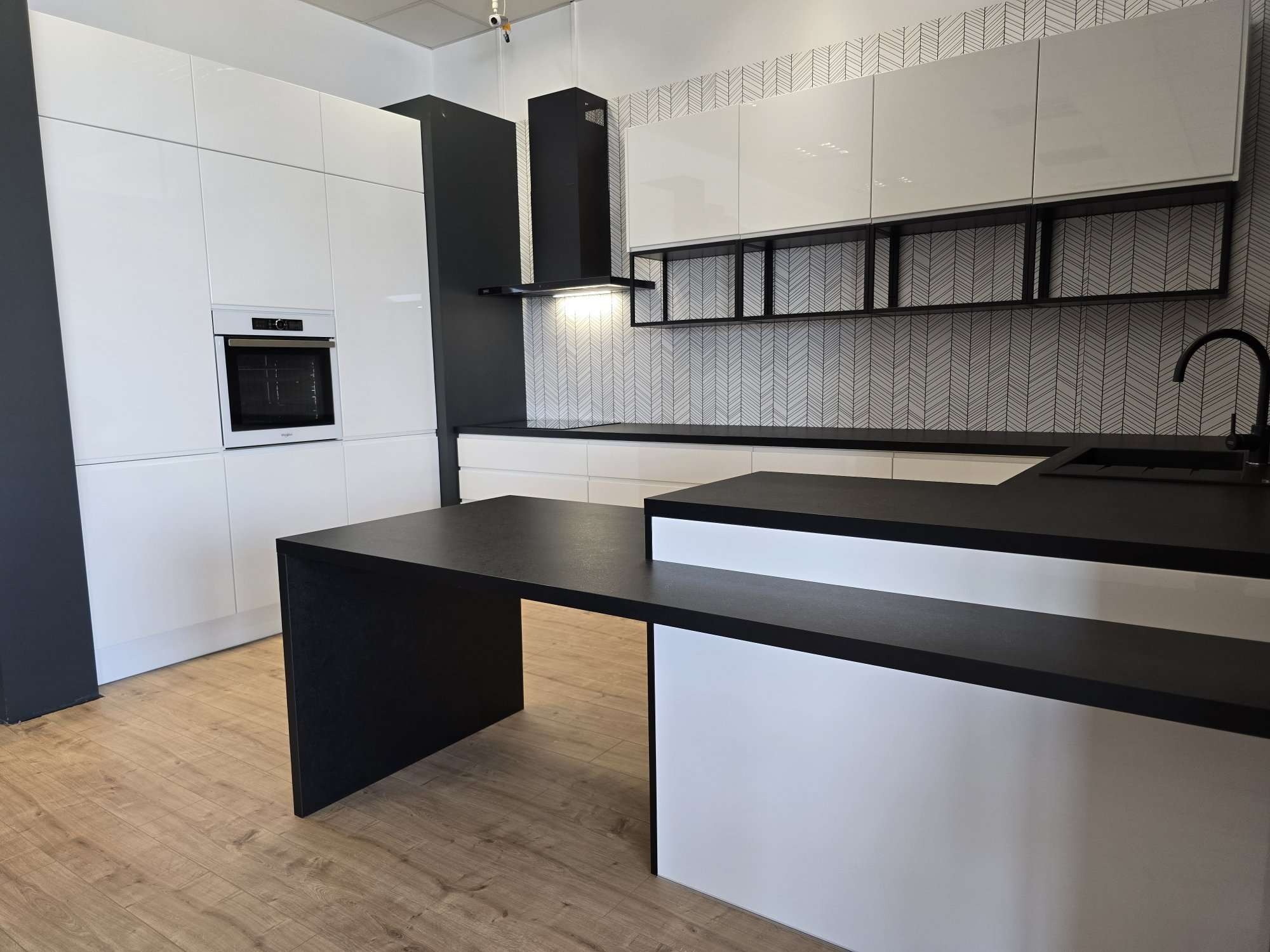 Nowoczesna kuchnia w bieli i czerni z wyspą, błyszczące fronty szafek górnych, czarny okap i blat, wzorzysta tapeta na ścianie.