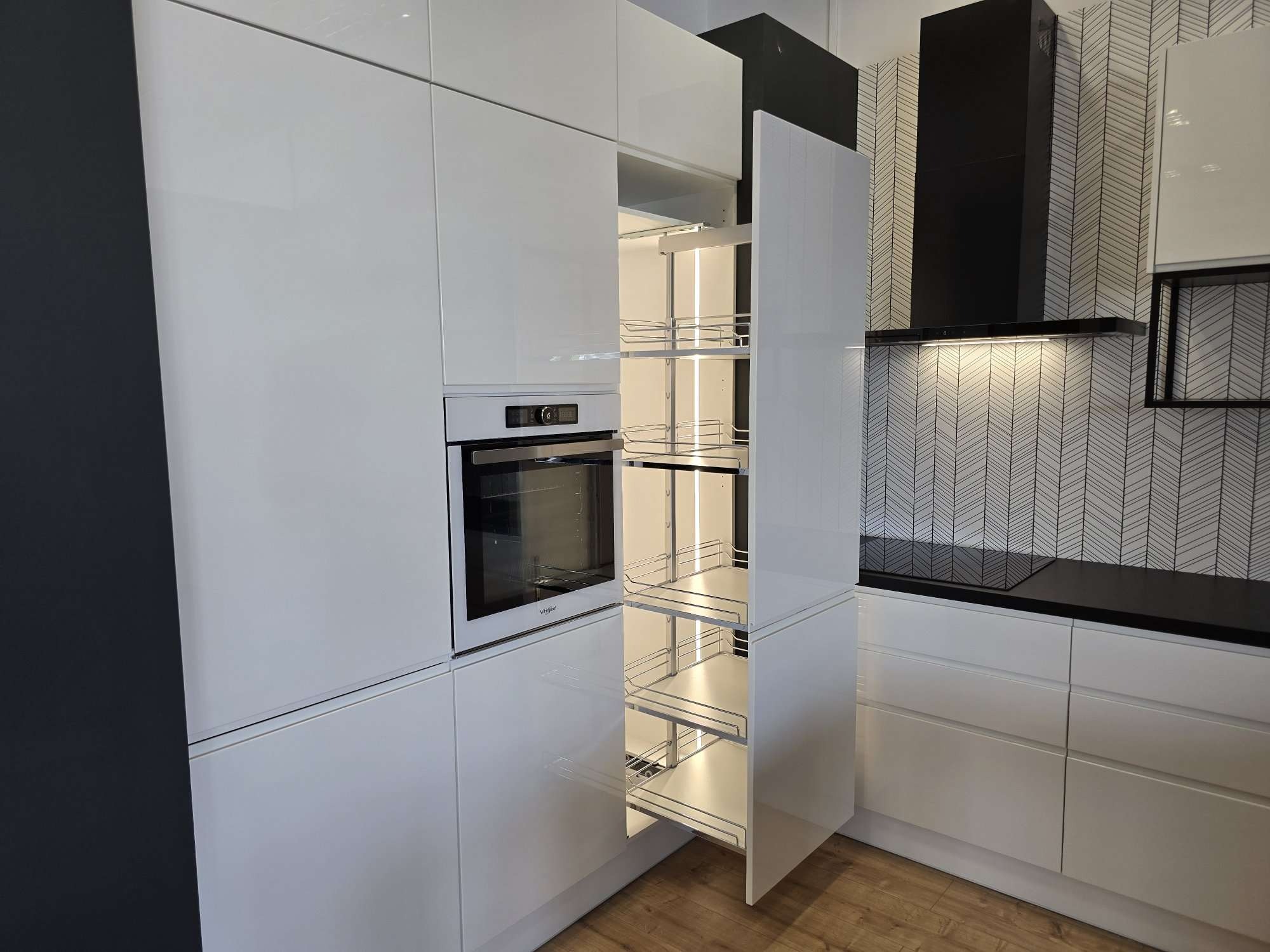 Biała, nowoczesna kuchnia z połyskiem, z wysuwaną szafką cargo z metalowymi koszami i wbudowanym piekarnikiem, na tle ściany z tapetą w geometryczny wzór.