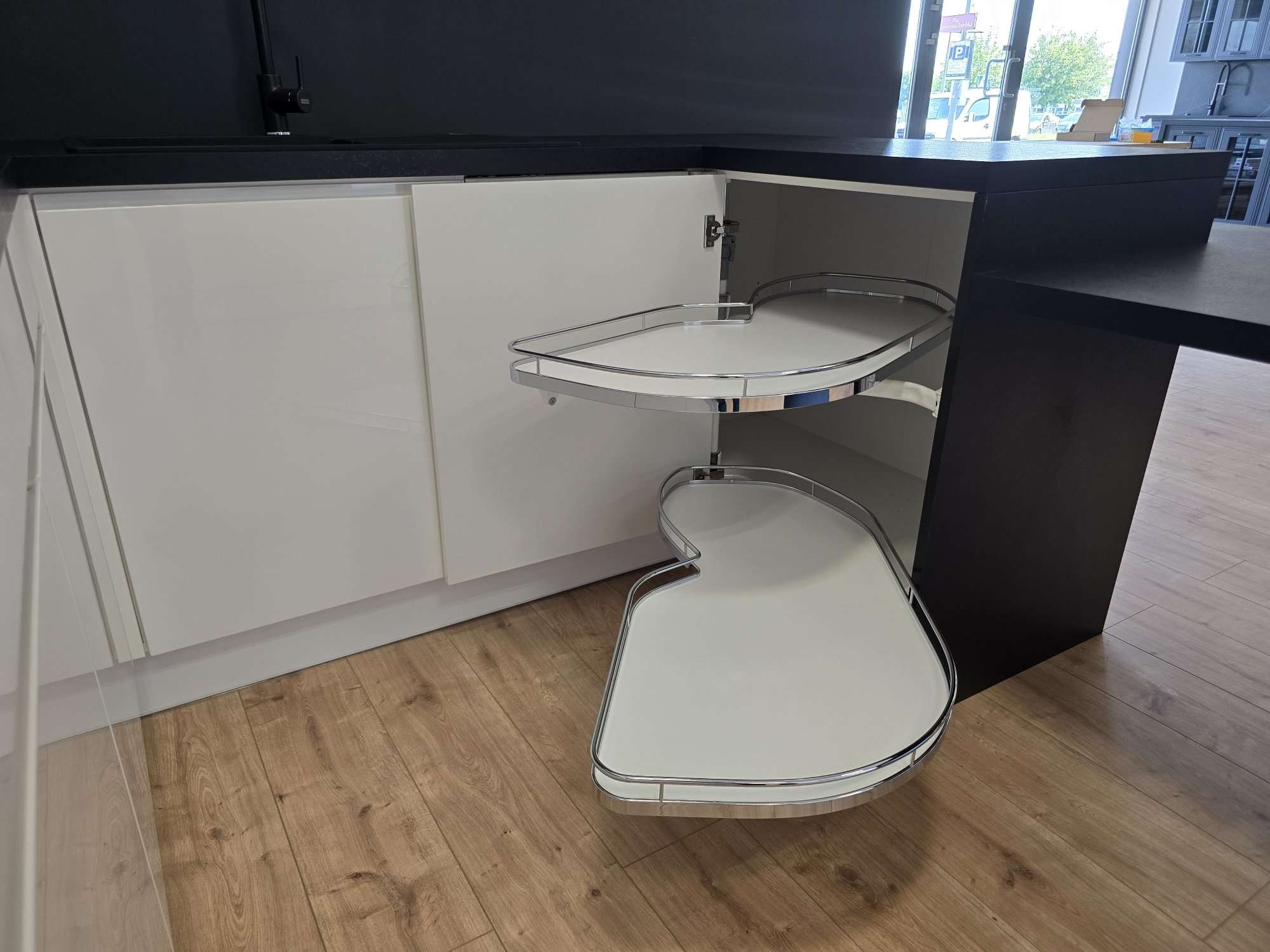 Wnętrze szafki narożnej z wysuwanym systemem półek obrotowych, widoczne białe fronty i ciemny blat, na podłodze panele imitujące drewno.