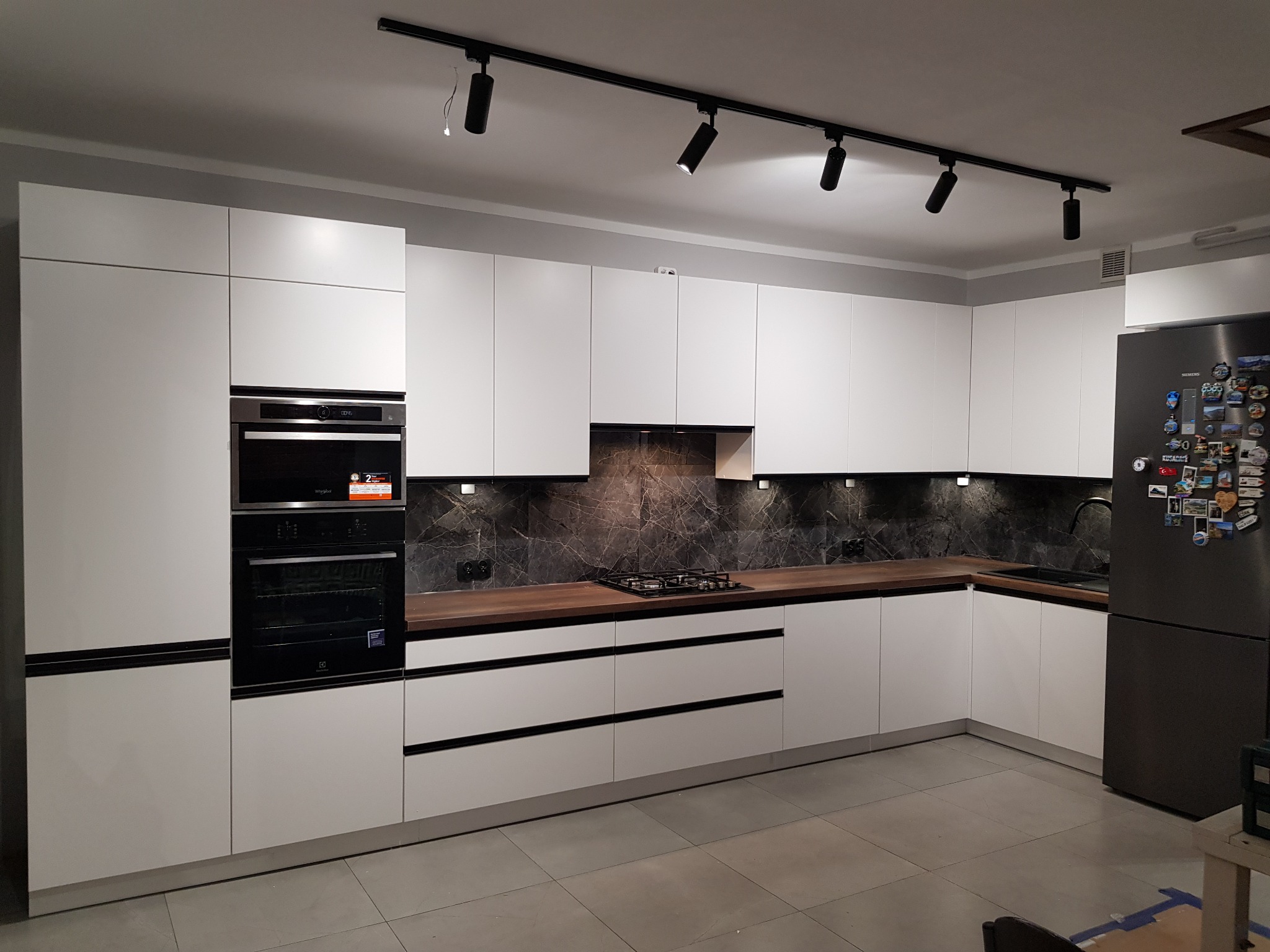 Nowoczesna, jasna kuchnia z białymi frontami szafek, drewnianym blatem i czarnym panelem nad blatem, oświetlona szynoprzewodem z reflektorami.