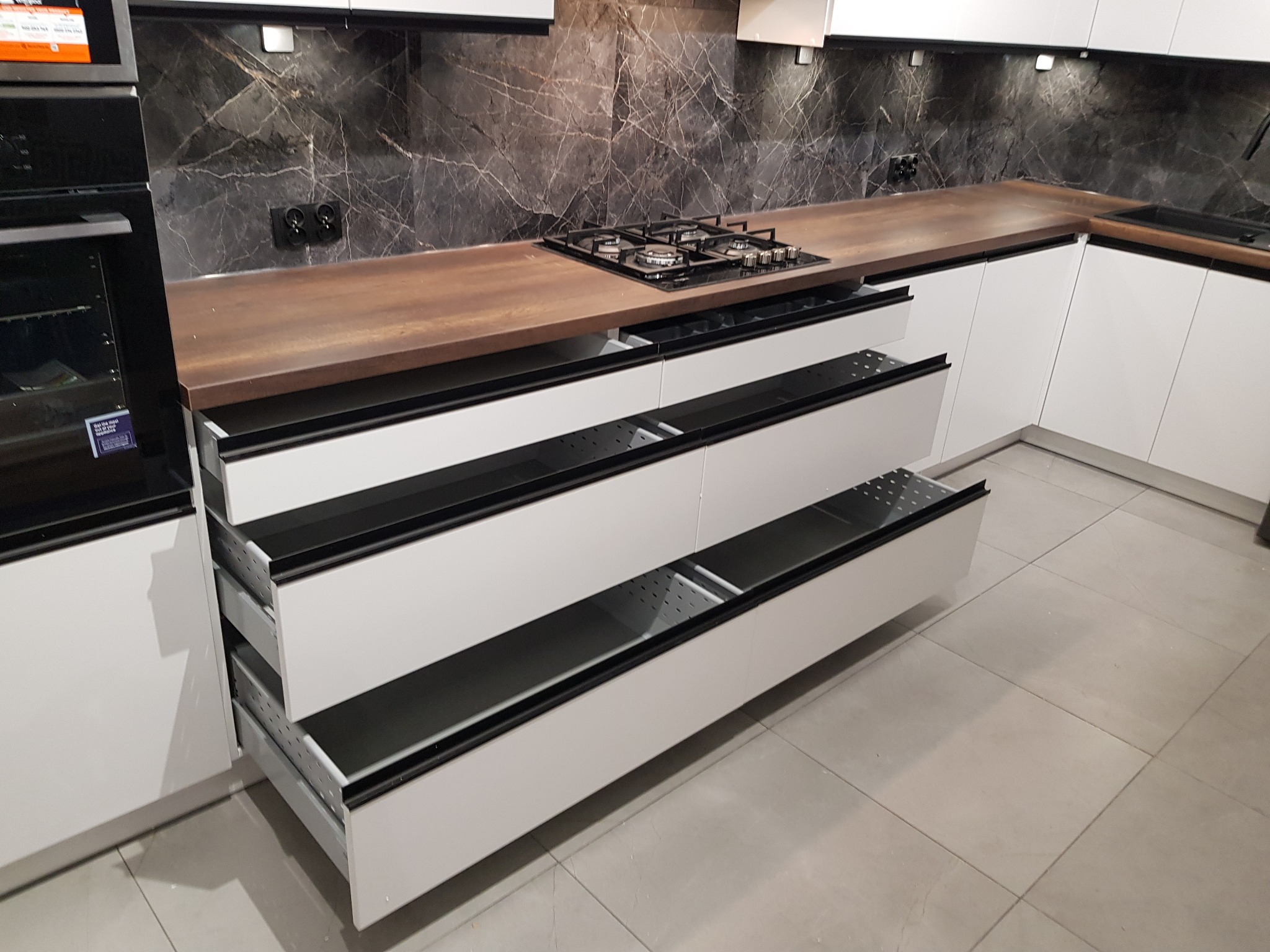 Nowoczesna kuchnia z białymi frontami, drewnianym blatem i otwartymi szufladami z perforowanym dnem, ukazującymi mechanizm wysuwania.