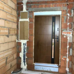 Montaż brązowych drzwi wejściowych z czarnym panelem w surowym murze z widoczną instalacją elektryczną i pianką montażową. Widoczne kable w peszlach.