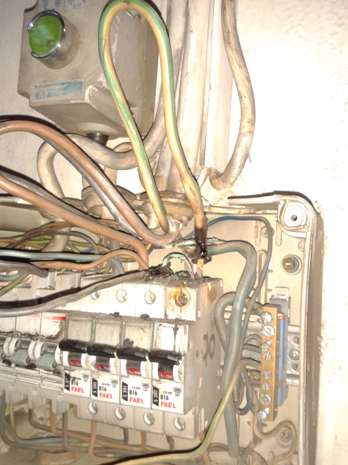 Uszkodzona skrzynka elektryczna z widocznymi przepalonymi przewodami i osmolonymi elementami. Bezpieczniki FAEL B16. Widoczne ślady zwarcia.