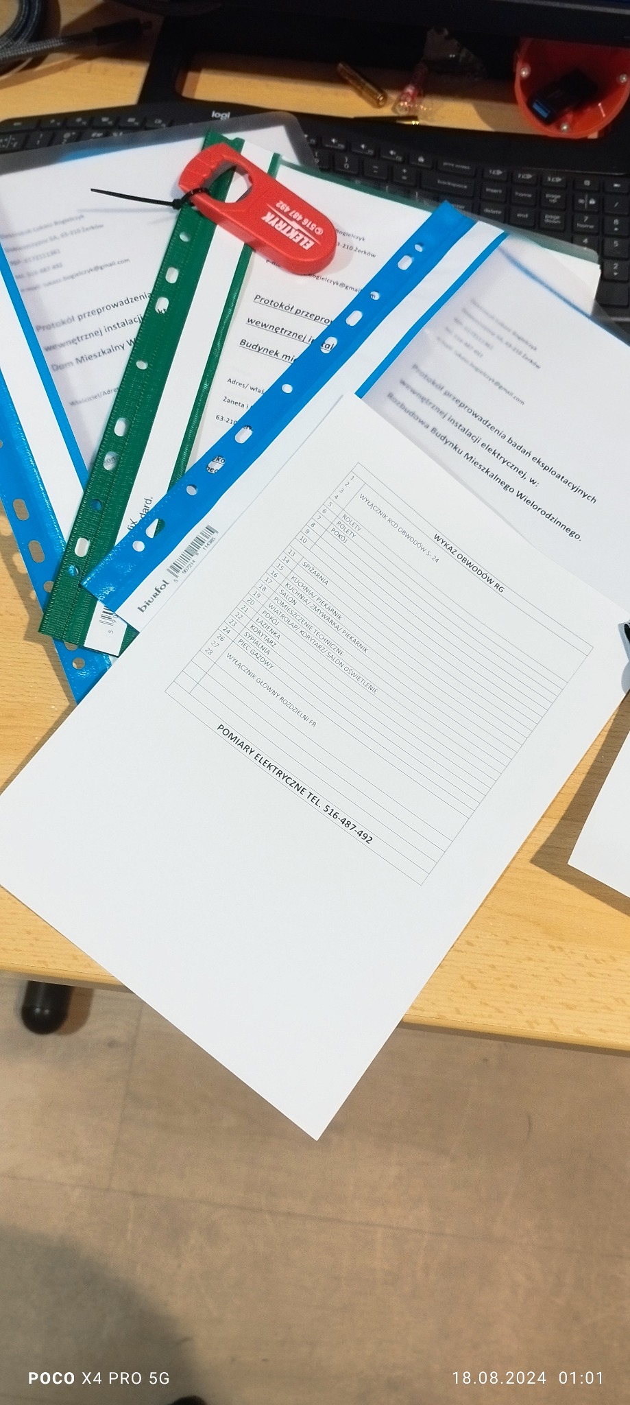 Stół z dokumentami dotyczącymi protokołów i pomiarów elektrycznych, w tym wykaz obwodów RG. Na wierzchu leży czerwony brelok z napisem 'Elektryk' i numerem telefonu.