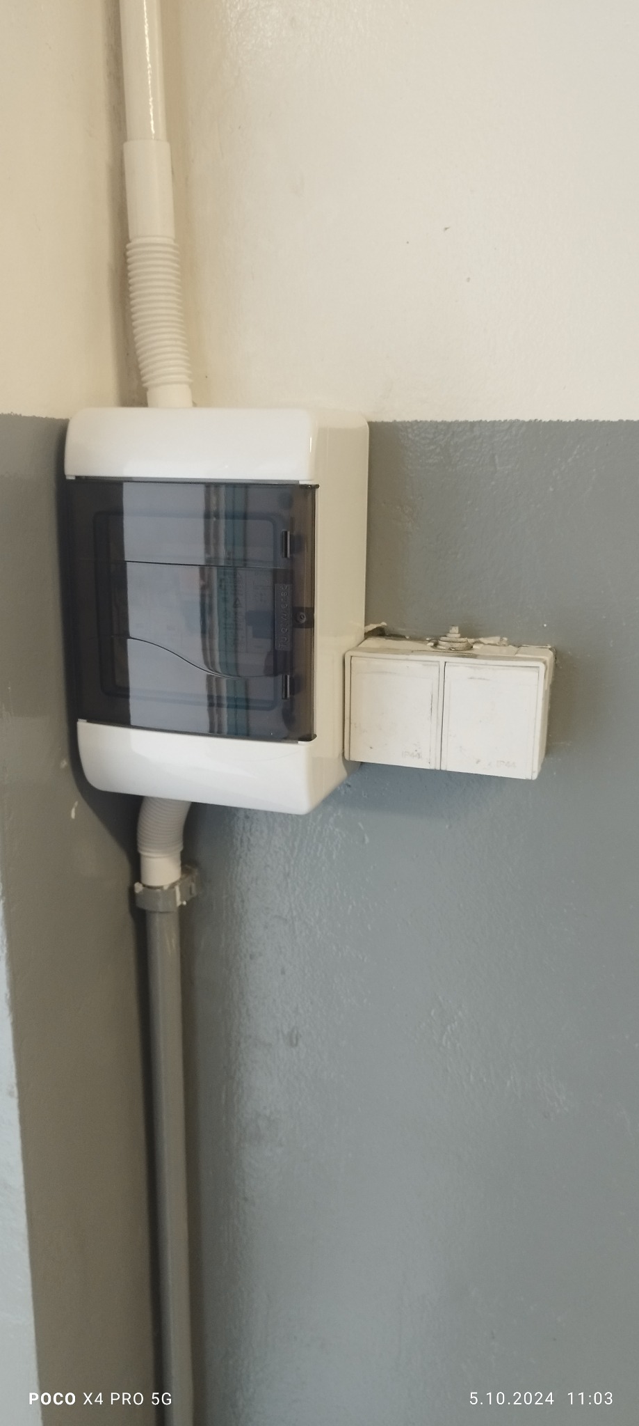 Zamknięta skrzynka elektryczna z plastikową osłoną i rurami ochronnymi przymocowana do szarej ściany obok podwójnego włącznika światła.