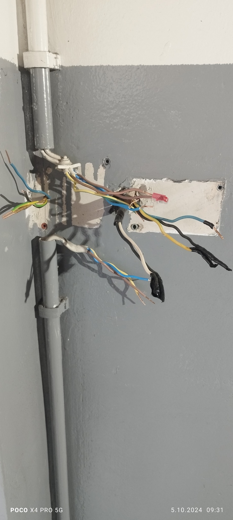 Odsłonięte przewody elektryczne wystające ze ściany pomalowanej na szaro, z widocznymi elementami instalacji w rurkach PCV.