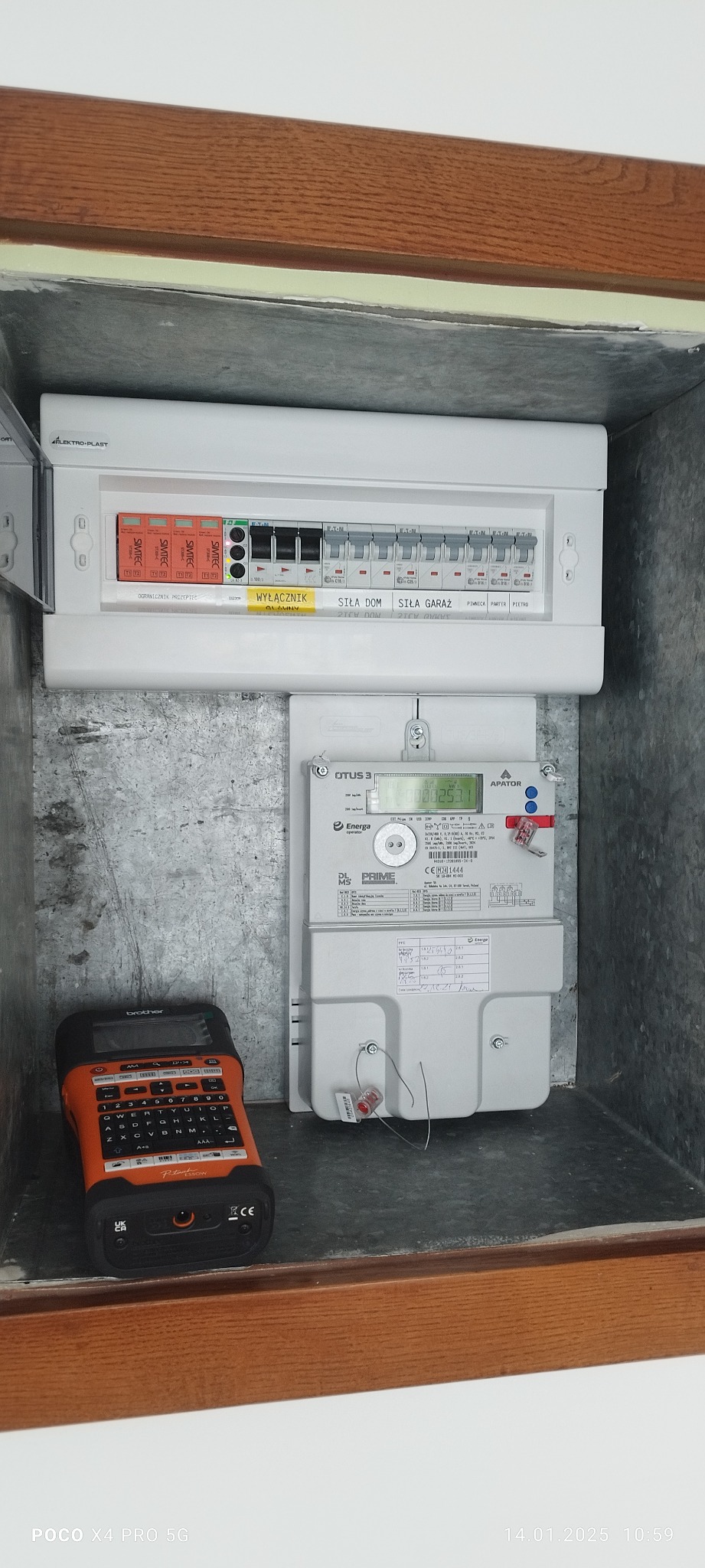 Skrzynka elektryczna z bezpiecznikami i licznikiem energii elektrycznej zamontowana w metalowej wnęce, obok leży etykieciarka Brother P-touch E550W.