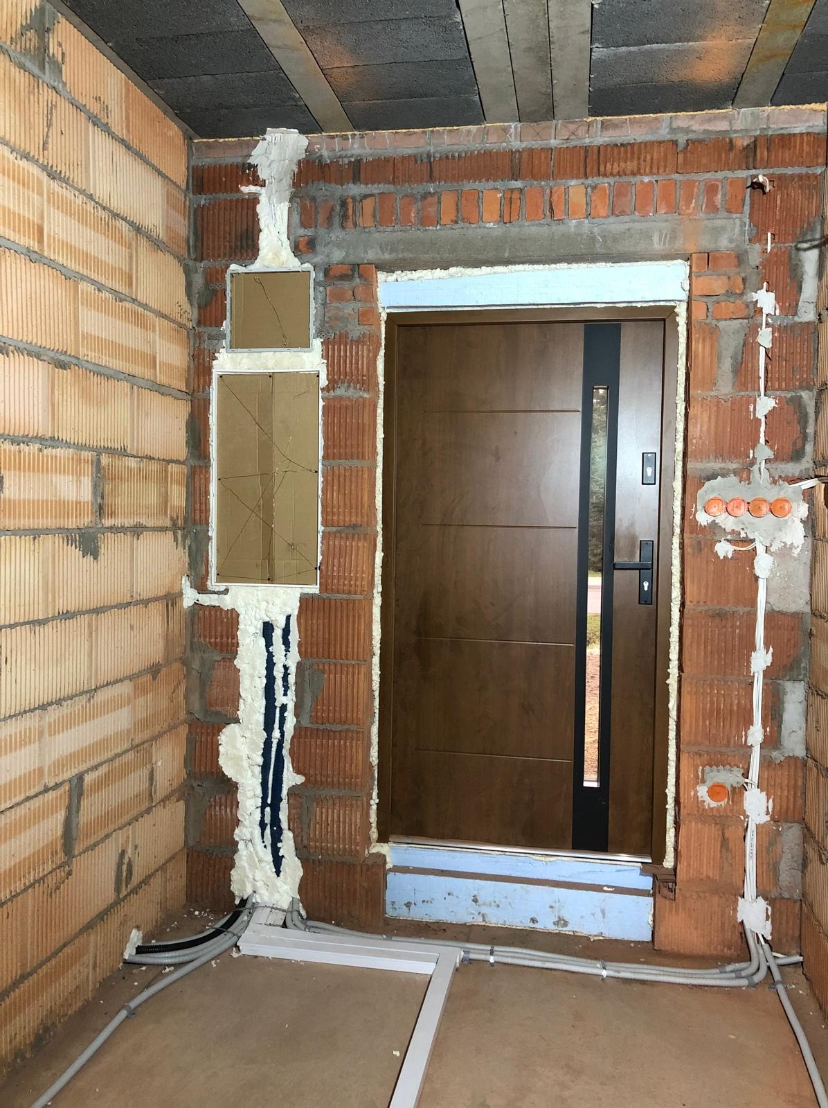 Montaż brązowych drzwi wejściowych z czarnym panelem w surowym murze z widoczną instalacją elektryczną i pianką montażową. Widoczne kable w peszlach.