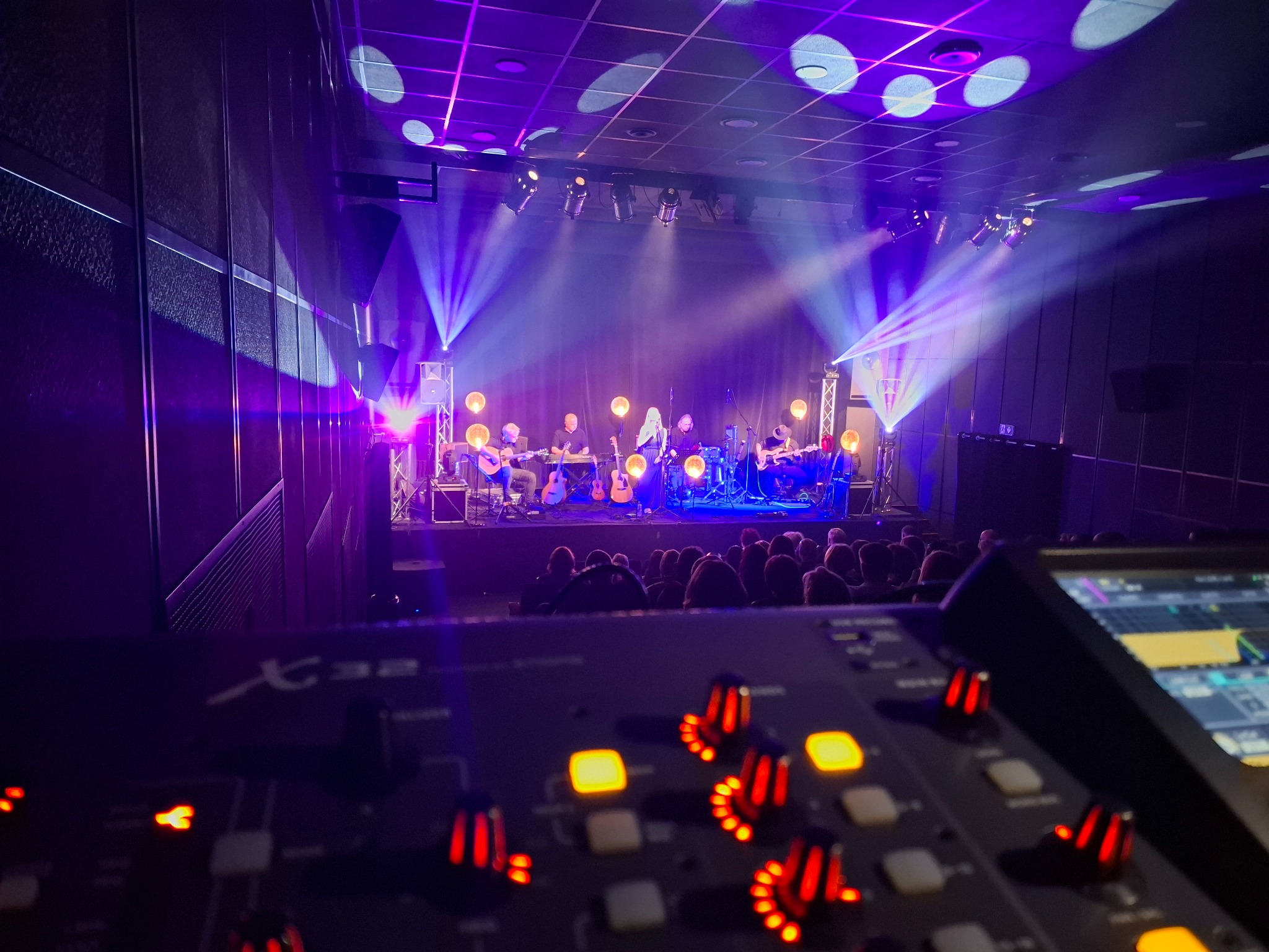 Zespół muzyczny na scenie oświetlonej fioletowymi reflektorami, widok zza konsoli mikserskiej z podświetlonymi przyciskami w kolorze czerwonym i żółtym.