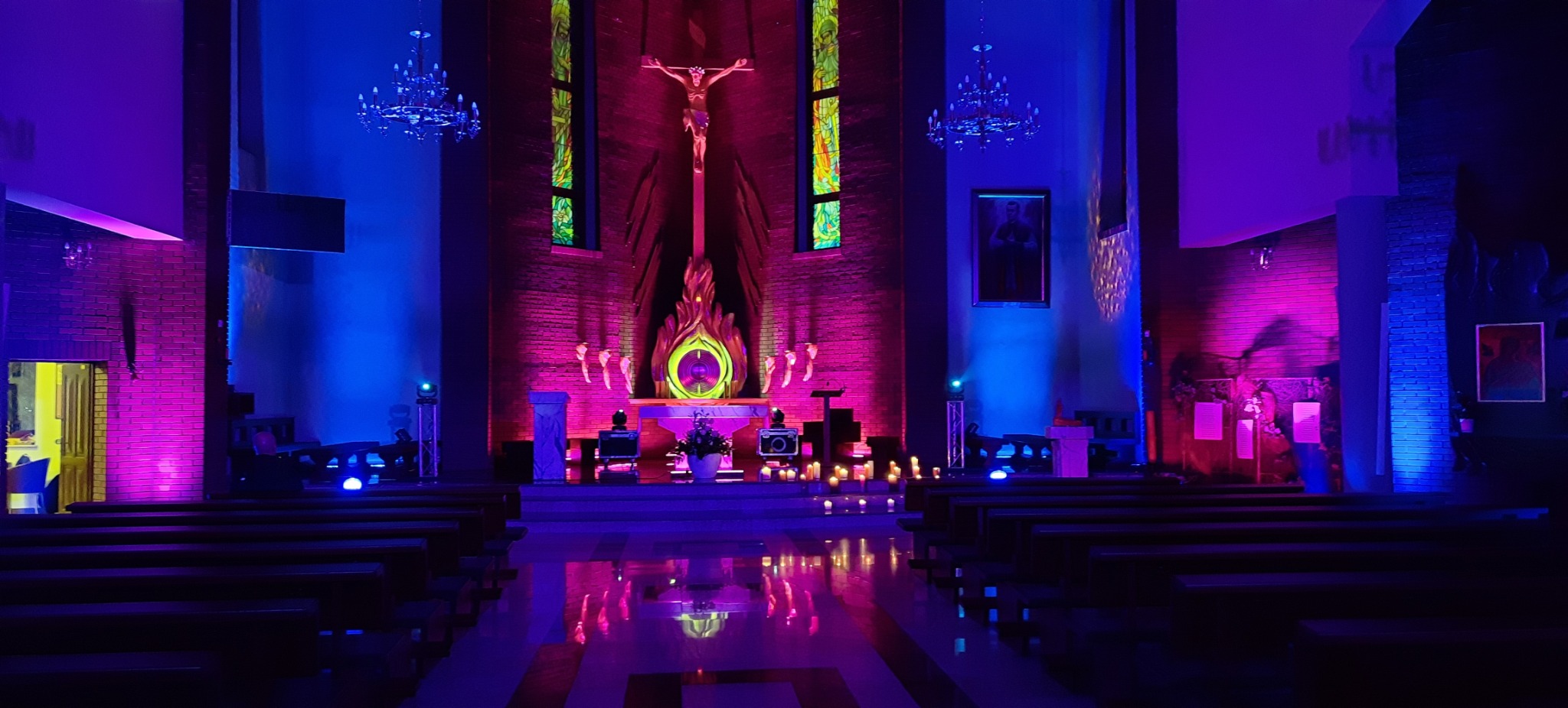 Wnętrze kościoła oświetlone intensywnym, dekoracyjnym światłem w odcieniach fioletu i niebieskiego, z reflektorami i świecami. Widoczny ołtarz i ławki.