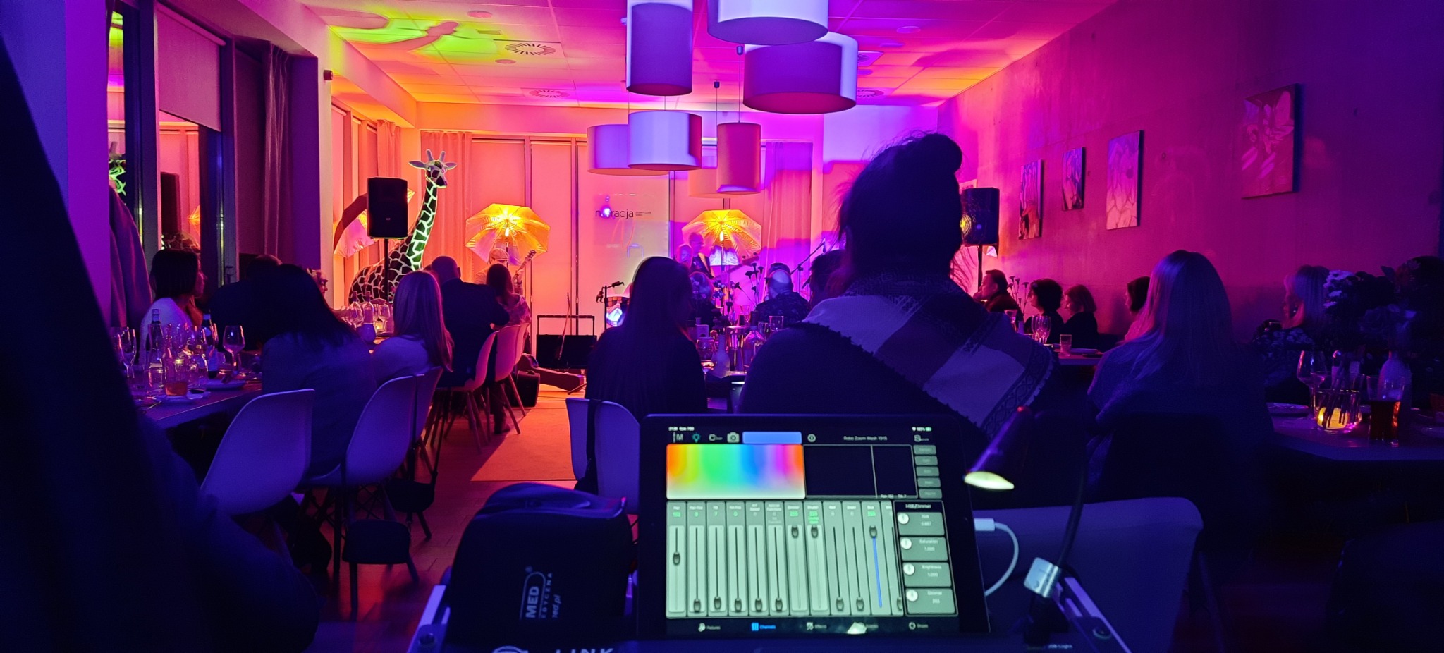 Kolorowe oświetlenie eventowe: sala z gośćmi, dekoracjami (żyrafa) i stołem mikserskim do sterowania światłem. Intensywne barwy, widok z perspektywy realizatora.