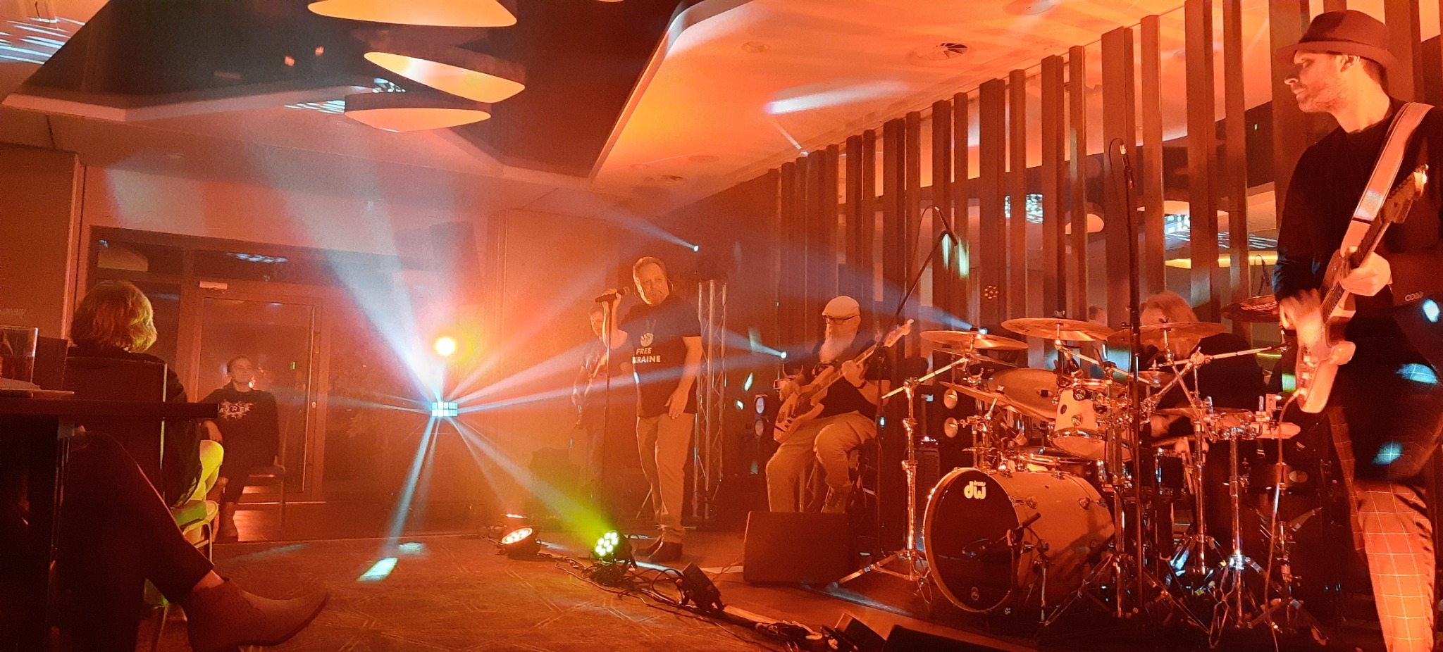 Zespół muzyczny na scenie z wokalistą w koszulce 'Free Ukraine', perkusją i gitarzystami, oświetlony reflektorami podczas występu na żywo.