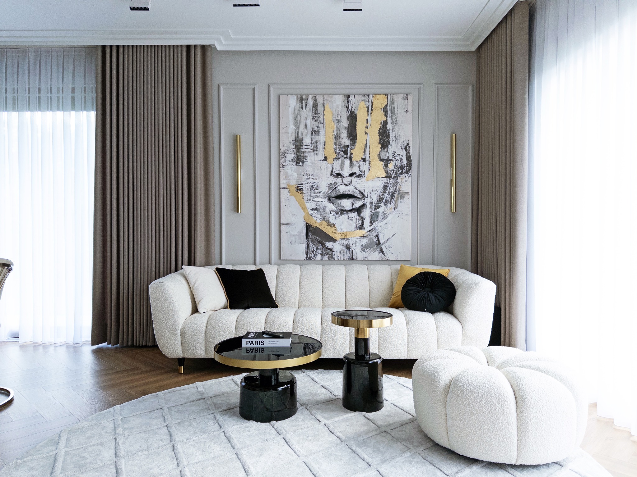 Elegancki salon w odcieniach szarości i bieli z akcentami złota, sofa z boucle, abstrakcyjny obraz z motywem twarzy i stoliki kawowe z czarnym blatem.