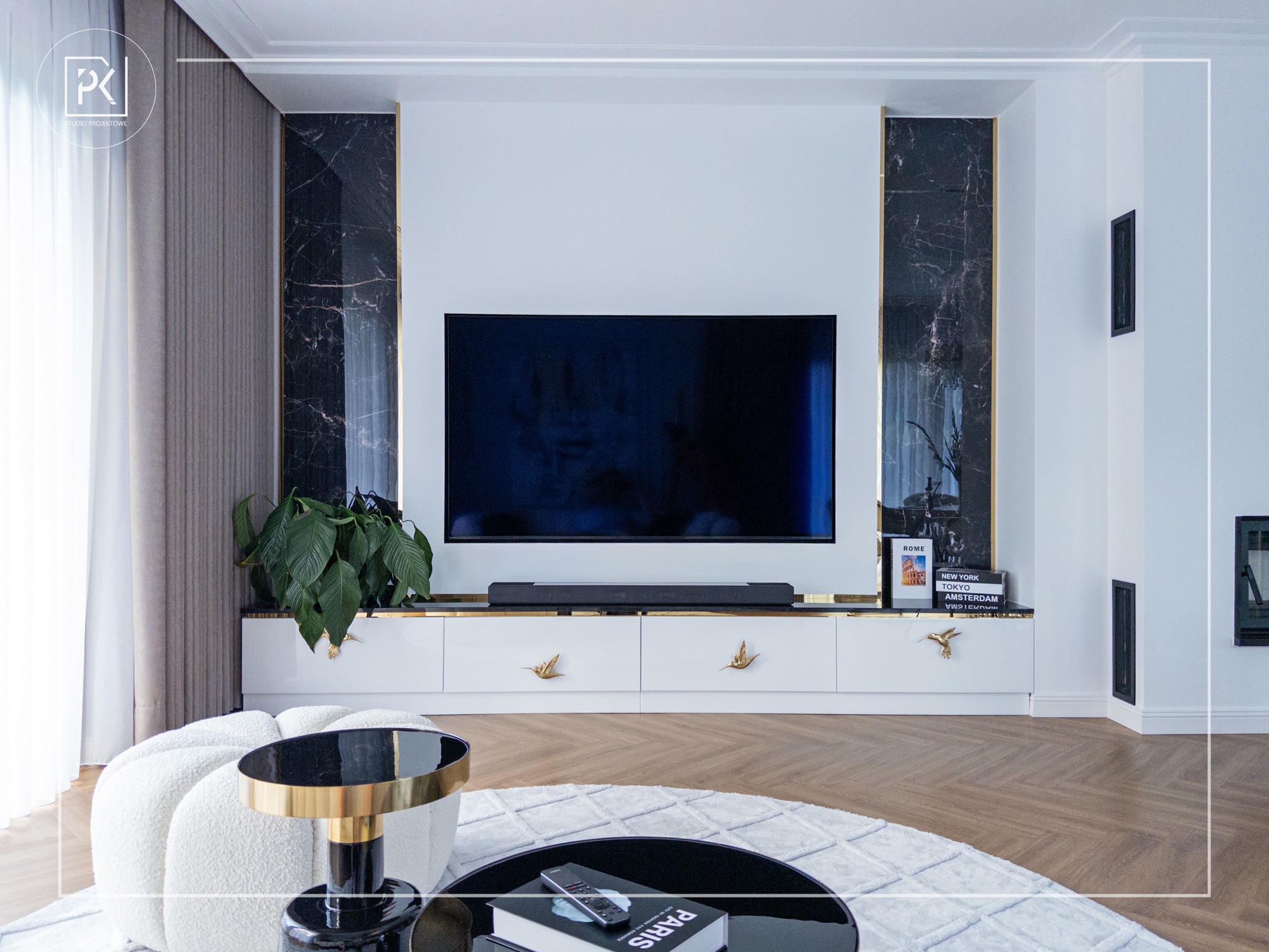 Elegancki salon z telewizorem na białej szafce RTV z dekoracyjnymi uchwytami w kształcie ptaków, marmurowe panele po bokach, okrągły stolik kawowy z czarnym blatem i złotymi detalami, puszysty...