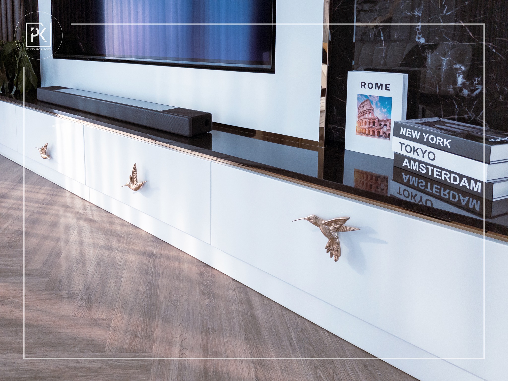 Biała szafka RTV z dekoracyjnymi uchwytami w kształcie kolibrów, nad nią czarny marmurowy blat z książkami o tematyce podróżniczej i soundbar pod telewizorem.