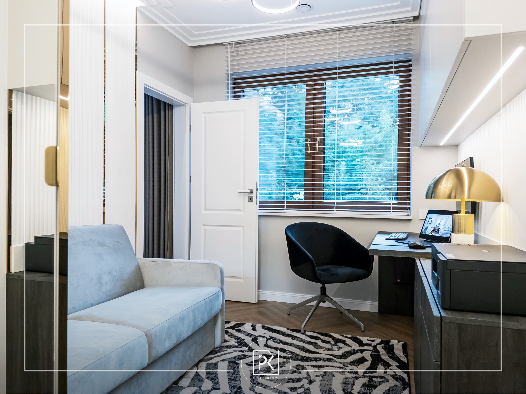 Nowoczesny gabinet z jasnoszarą sofą, czarnym krzesłem obrotowym, biurkiem z laptopem i drukarką, dywanem w zebrę oraz oknem z żaluzjami, oświetlony złotą lampką biurkową.