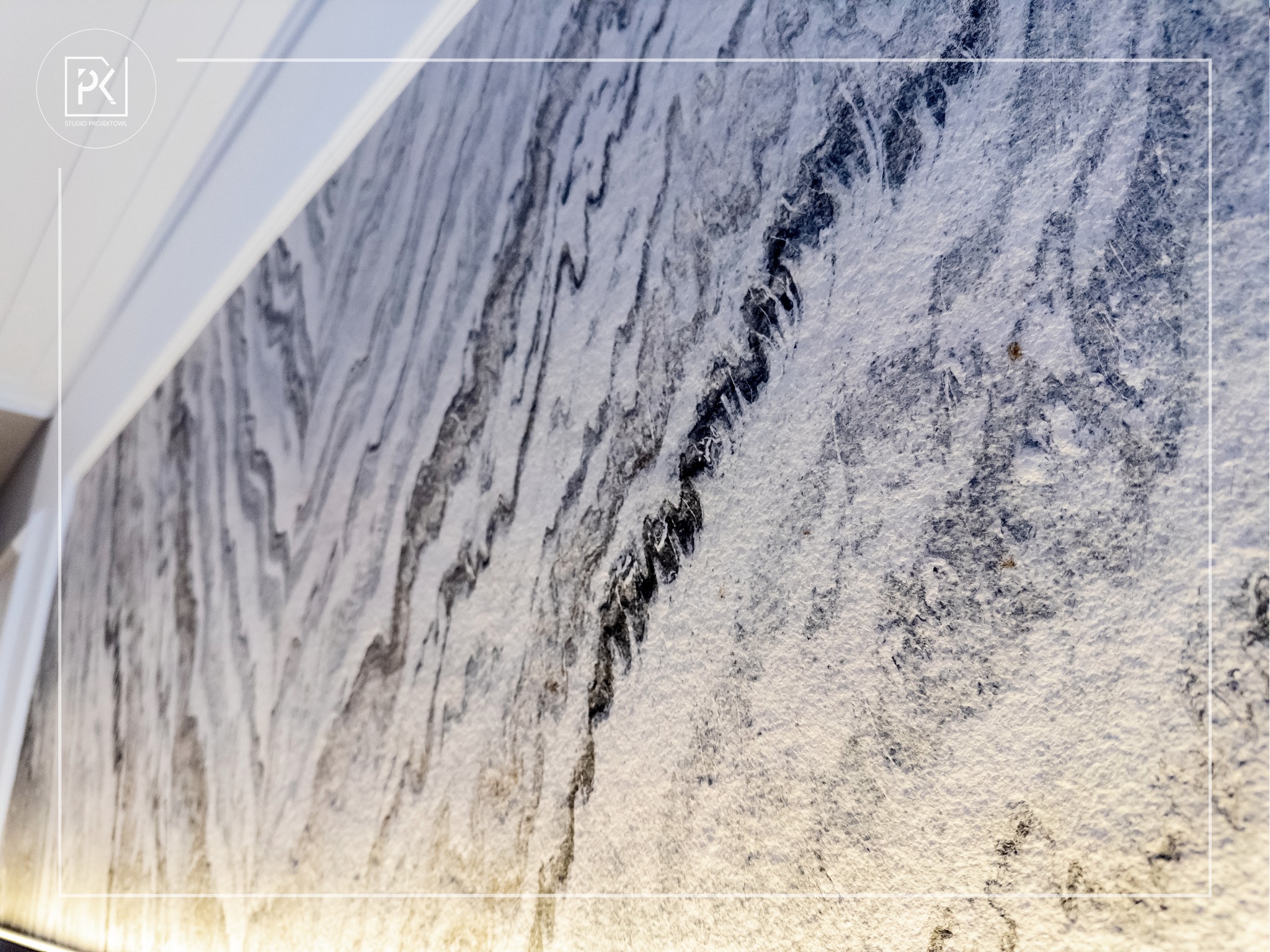 Fragment ściany z dekoracyjną tapetą o wzorze imitującym marmur w odcieniach szarości i bieli, oświetlony od dołu ciepłym światłem, z widocznym logo studia projektowego w lewym górnym rogu.