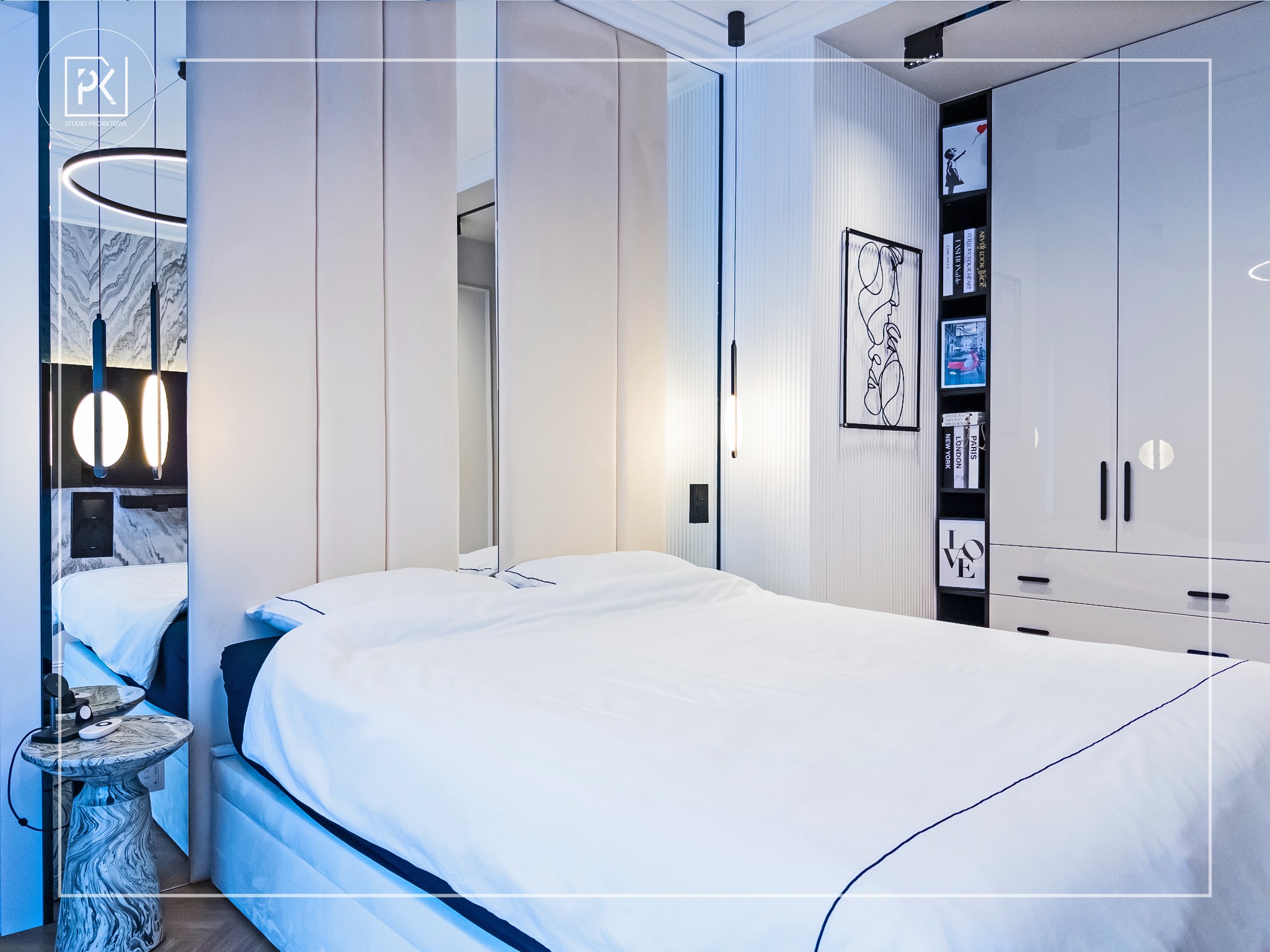 Nowoczesna sypialnia z łóżkiem w białej pościeli z granatowym obszyciem, marmurowy stolik nocny, lustrzane panele ścienne i zabudowa meblowa z półkami na książki i dekoracje.