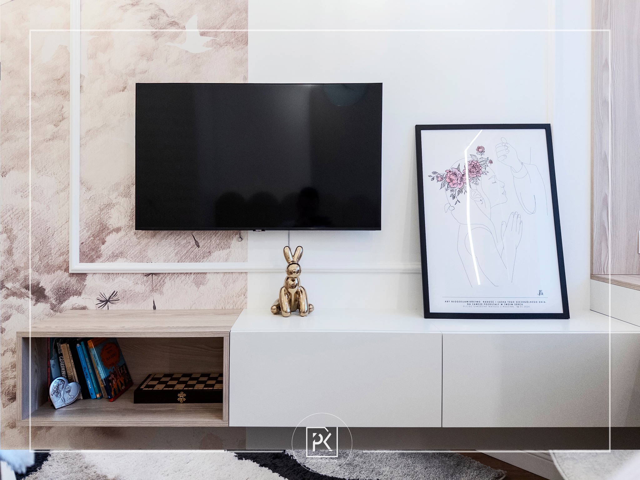Nowoczesny salon z telewizorem, białą szafką RTV, złotą figurką królika i abstrakcyjnym plakatem w czarnej ramie. Na ścianie wzorzysta tapeta w odcieniach beżu i brązu.