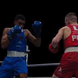 Aureliusz Brzostkiewicz - Dwóch bokserów na ringu: jeden w niebieskim stroju z godłem Niemiec, drugi w czerwonym z napisem POL, obaj w rękawicach bokserskich, w trakcie walki.