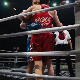 Aureliusz Brzostkiewicz - Dwóch bokserów obejmuje się w ringu po walce, widoczny sędzia w tle, sznurki ringu w pierwszym planie.