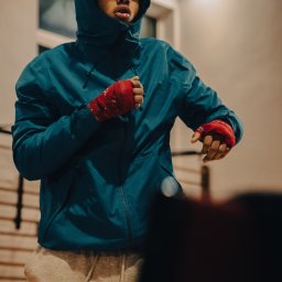 Aureliusz Brzostkiewicz - Młody mężczyzna w niebieskiej kurtce z kapturem i czerwonych bandażach na dłoniach, w trakcie treningu bokserskiego, patrzący w górę.