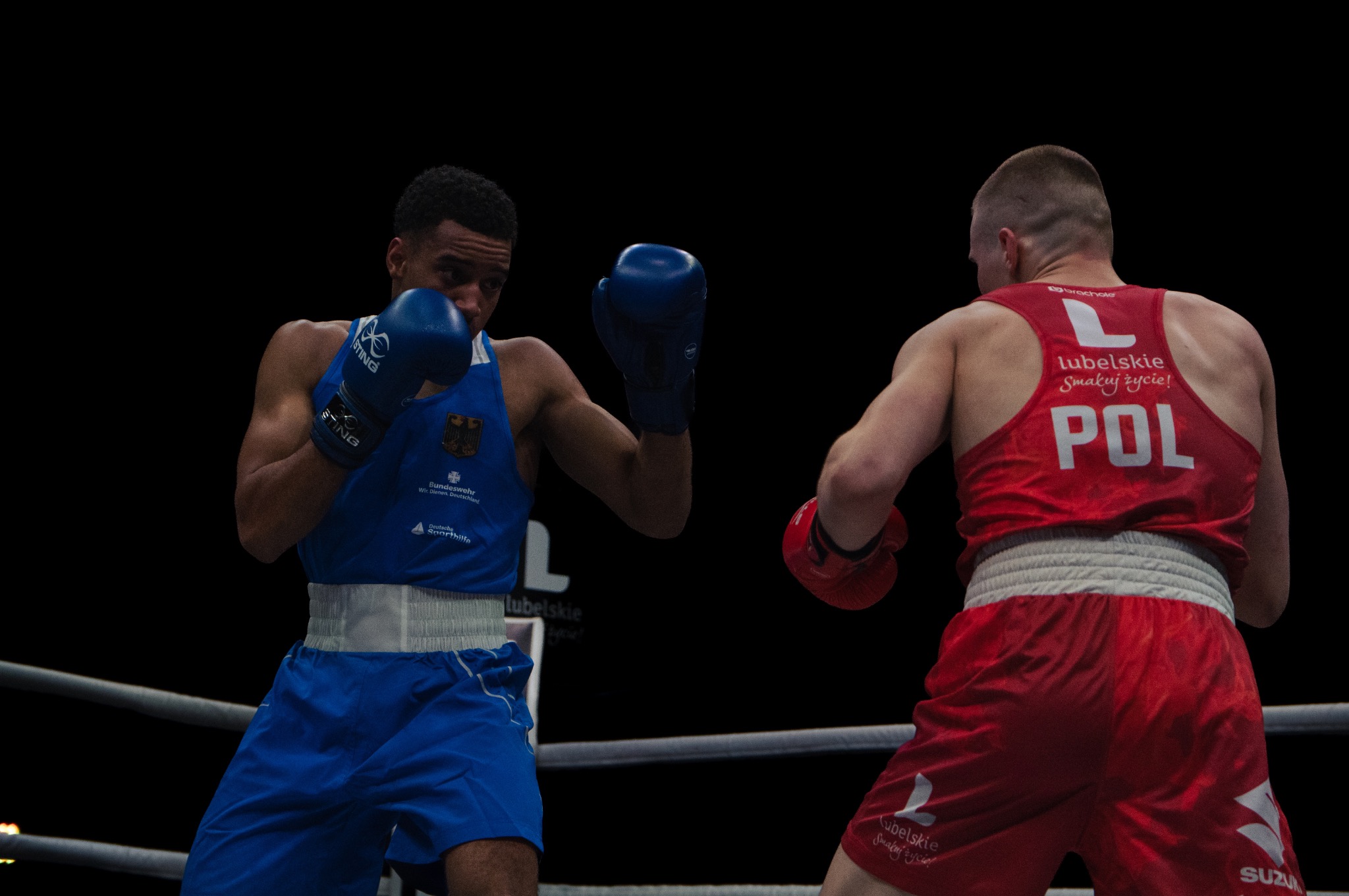 Dwóch bokserów na ringu: jeden w niebieskim stroju z godłem Niemiec, drugi w czerwonym z napisem POL, obaj w rękawicach bokserskich, w trakcie walki.