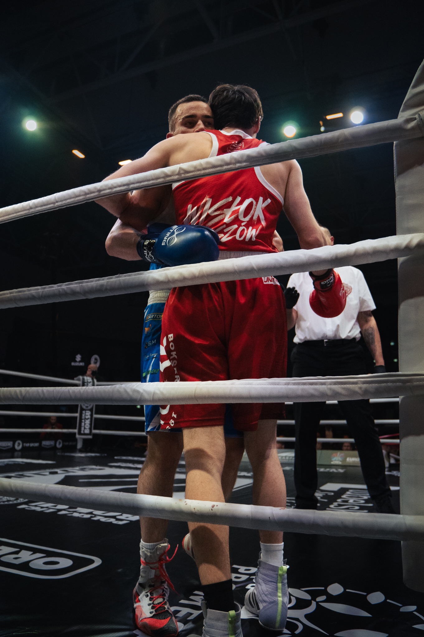 Dwóch bokserów obejmuje się w ringu po walce, widoczny sędzia w tle, sznurki ringu w pierwszym planie.