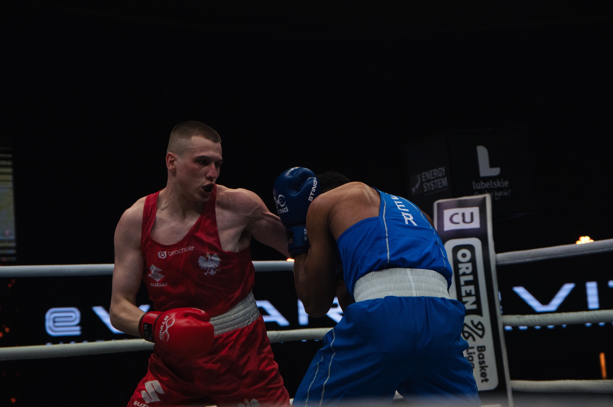 Dwóch bokserów w ringu podczas zawodów: zawodnik w czerwonym stroju z godłem Polski wyprowadza cios w kierunku przeciwnika w niebieskim stroju, widoczne logotypy sponsorów na bandach ringu.