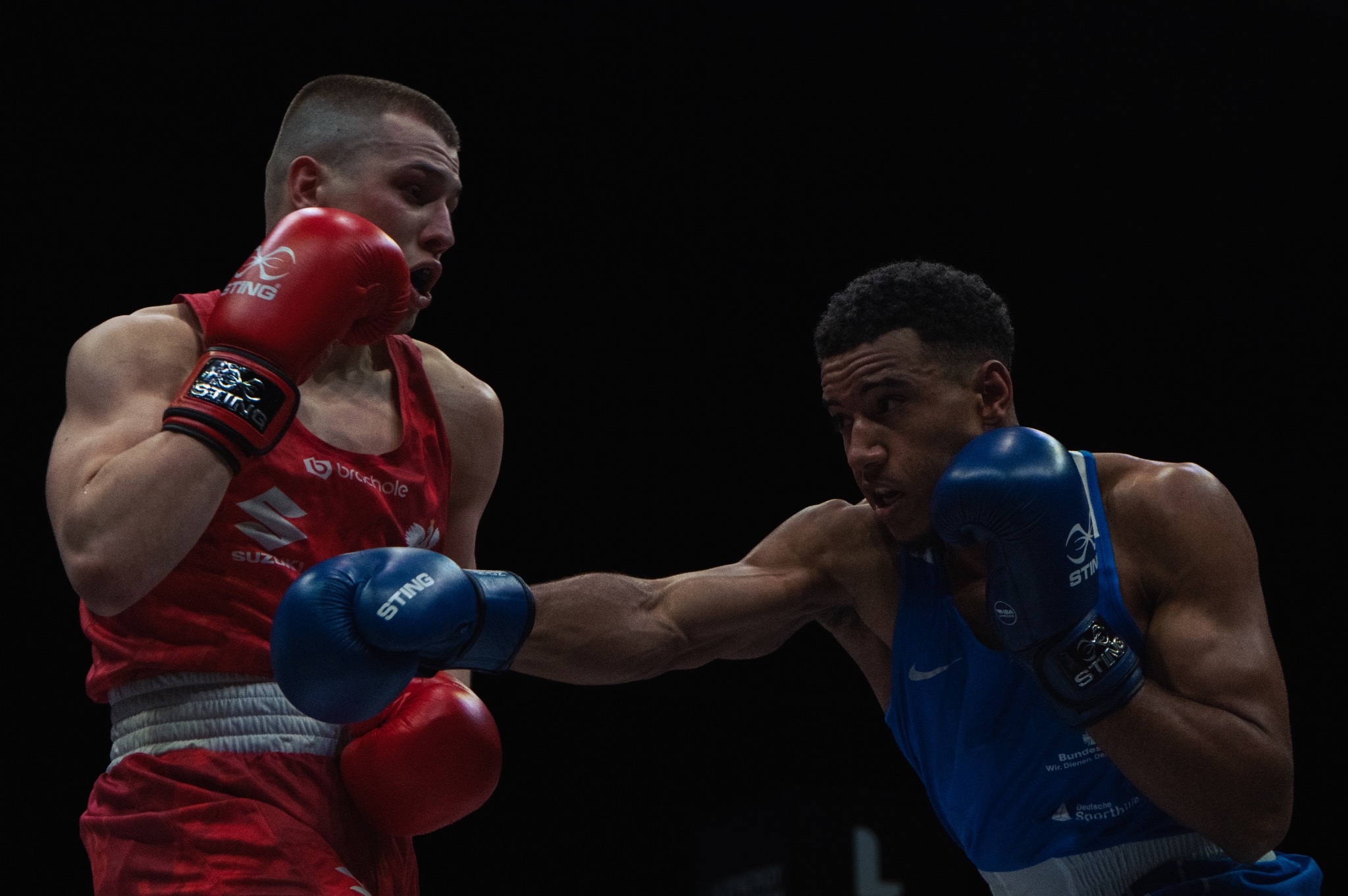 Dwóch bokserów w akcji: jeden w czerwonym stroju otrzymuje cios w twarz od przeciwnika w niebieskim stroju na czarnym tle.