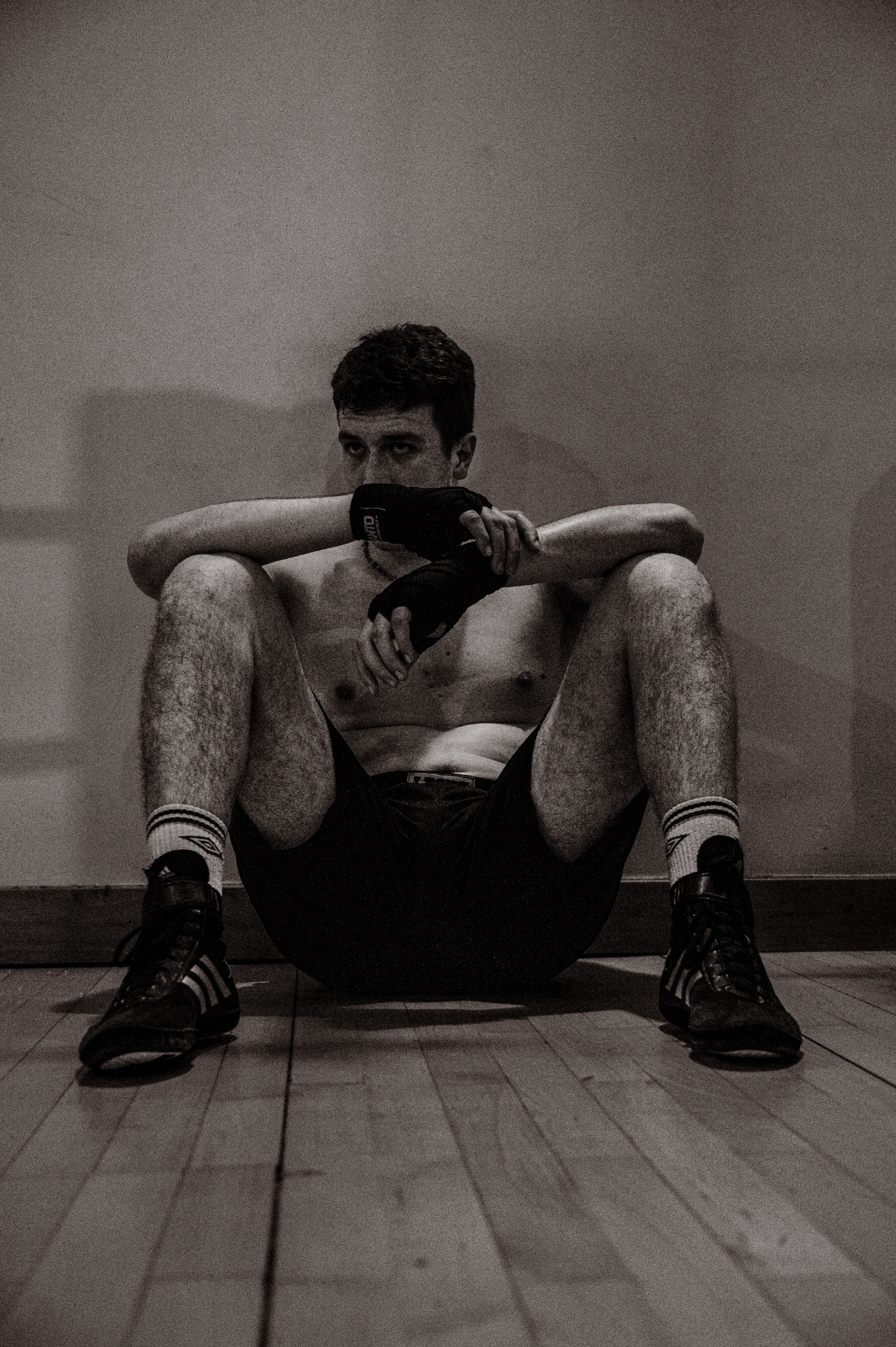 Czarno-białe zdjęcie siedzącego na podłodze mężczyzny po treningu, z bandażami na rękach, w sportowych butach Adidas i wysokich skarpetach.