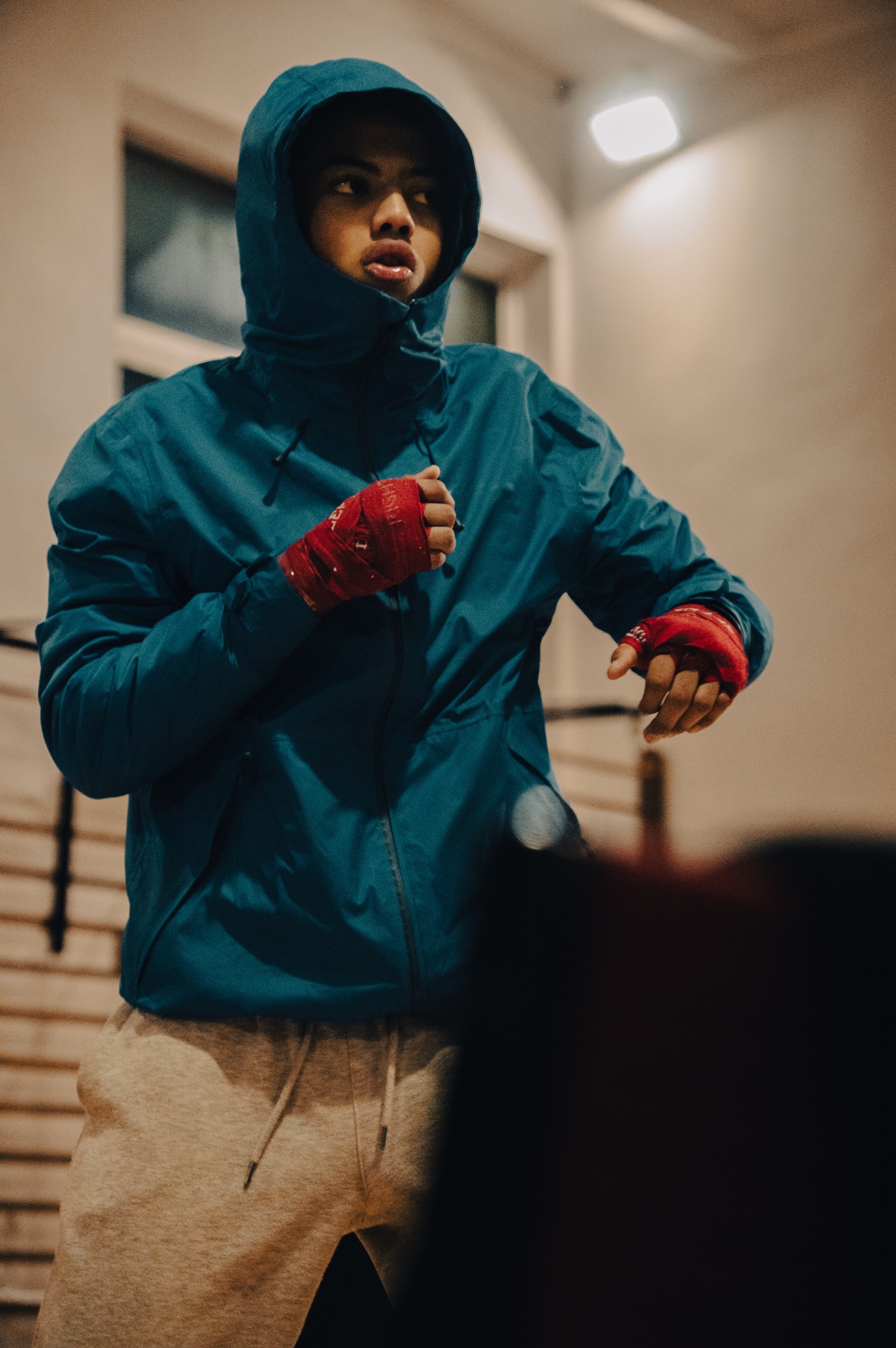 Młody mężczyzna w niebieskiej kurtce z kapturem i czerwonych bandażach na dłoniach, w trakcie treningu bokserskiego, patrzący w górę.