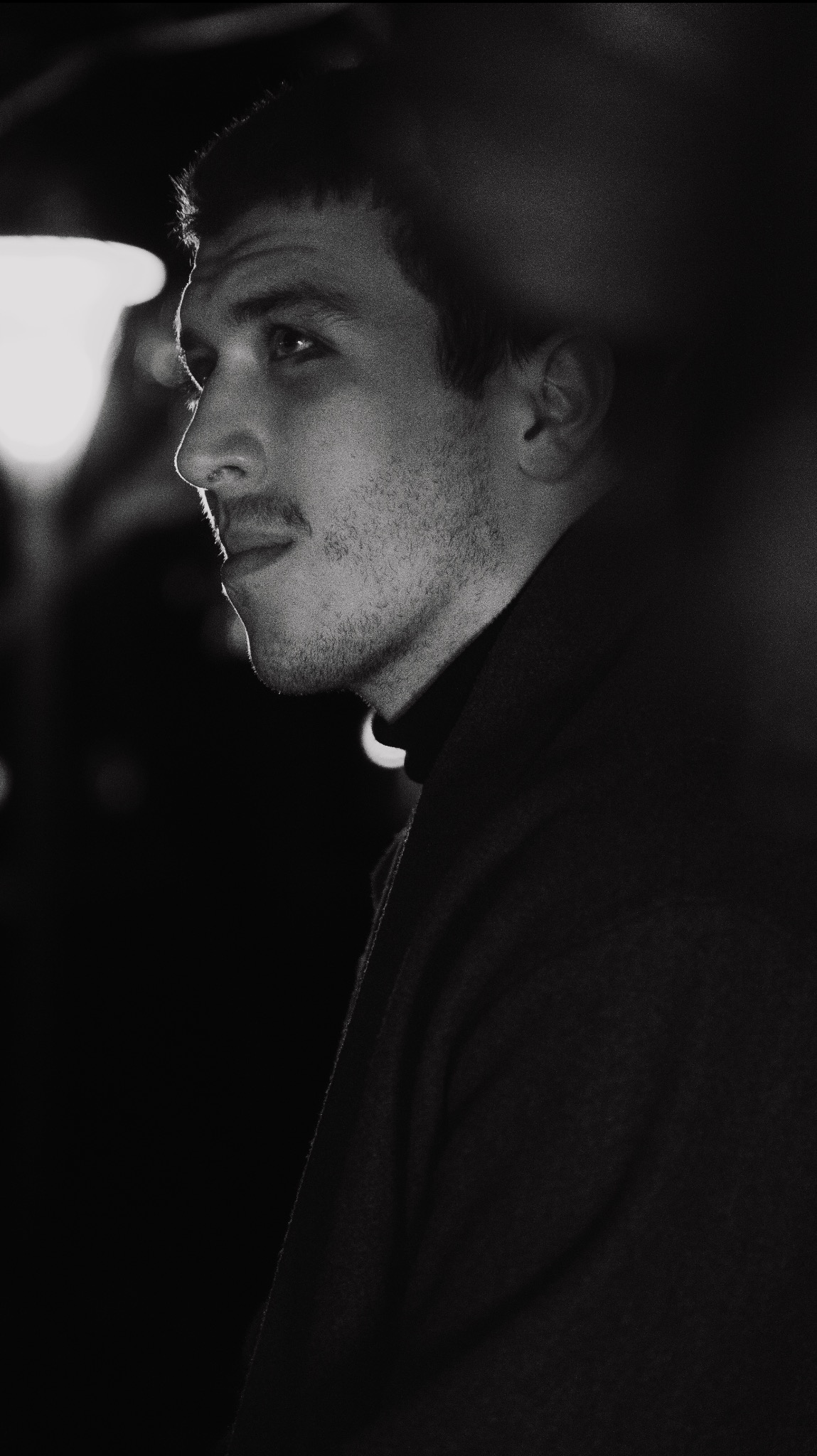 Czarno-białe portretowe ujęcie z profilu młodego mężczyzny w czarnym golfie, oświetlonego bocznym światłem, z widocznym fragmentem lampy w tle.